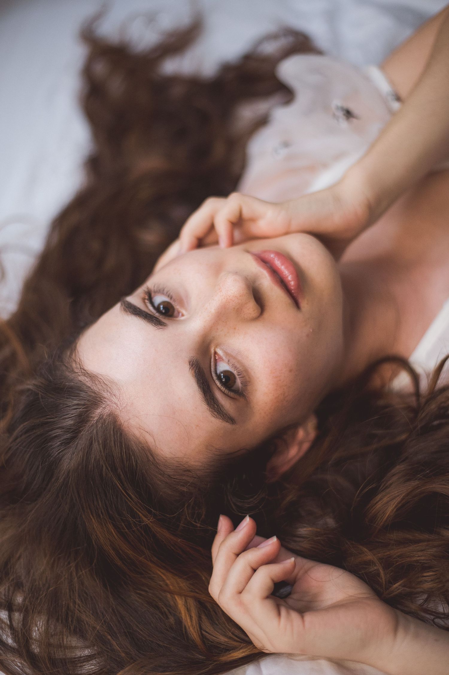 Young woman with long wavy brown hair lying on white bed, gazing upward with hands near her face.