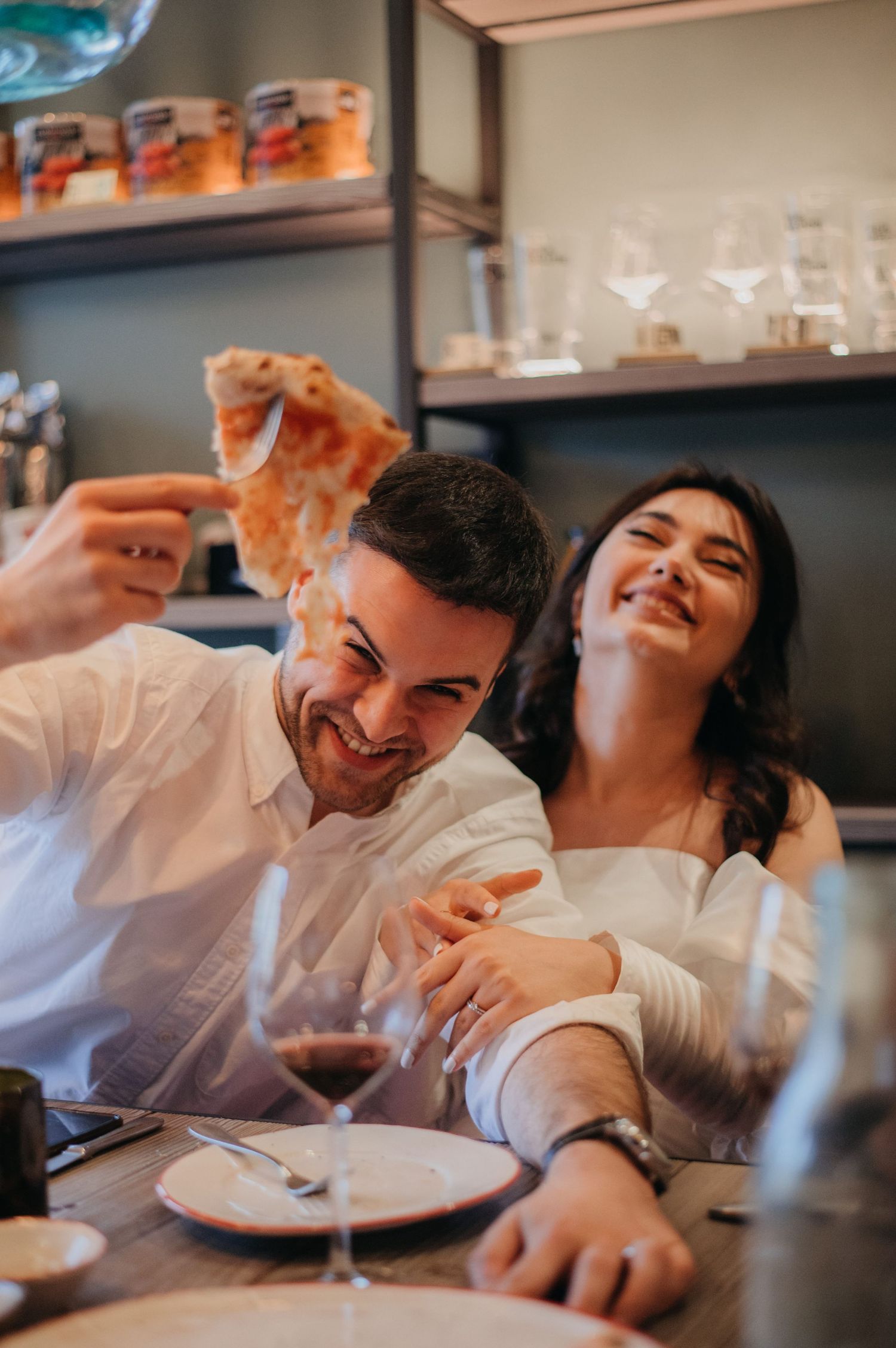 Happy couple sharing pizza slice and red wine at cozy restaurant table after wedding.