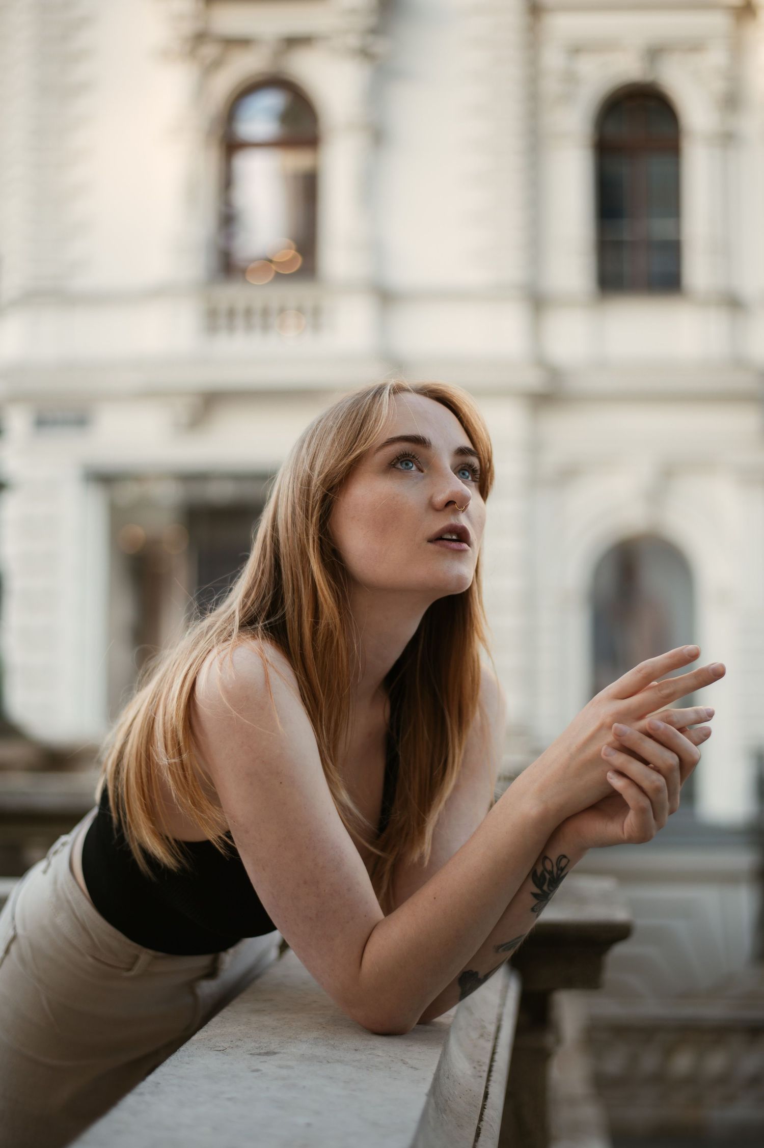 Young woman with long blonde hair leans on stone balustrade gazing upward at classic European building.