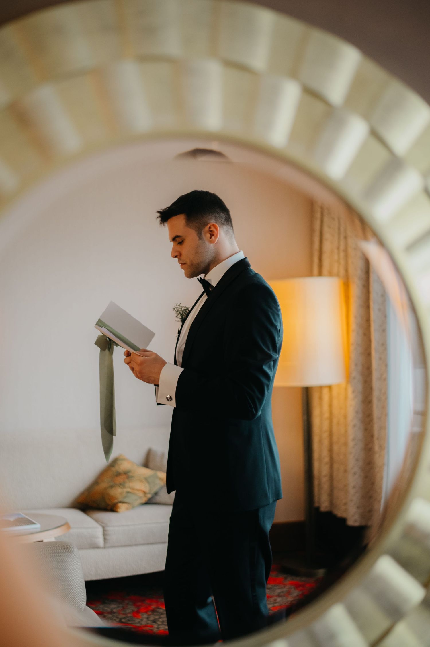 Groom in black tuxedo reading a letter reflected in decorative round mirror in hotel room.