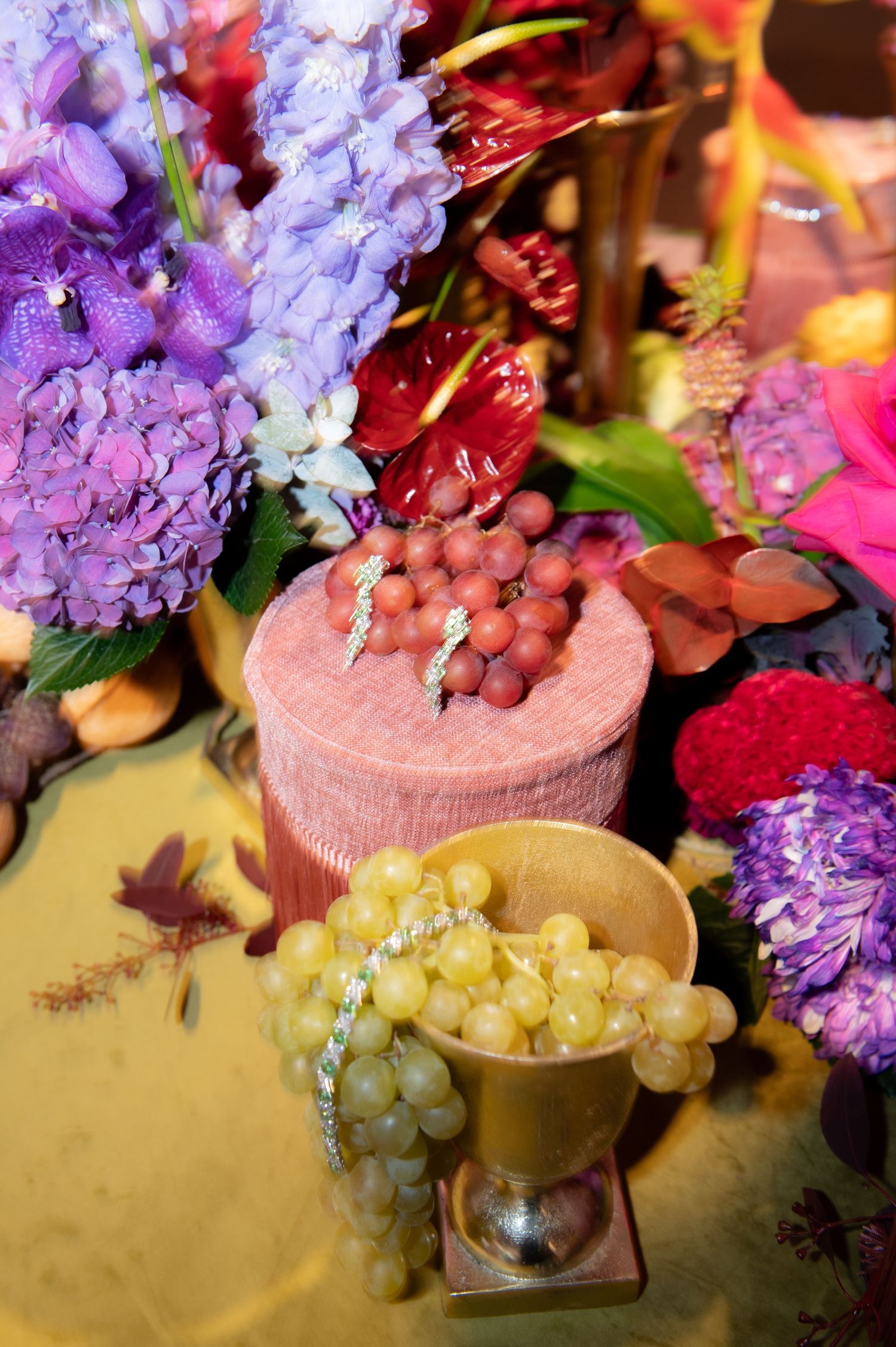 Colorful floral arrangement with purple hydrangeas, red anthuriums, and fresh green and red grapes in a gold cup.