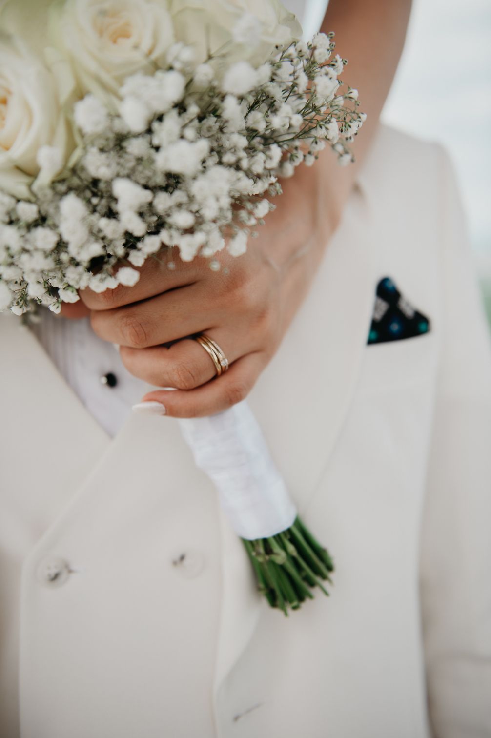 Close-up of bride holding a white rose and baby's breath bouquet with gold wedding rings on her finger.