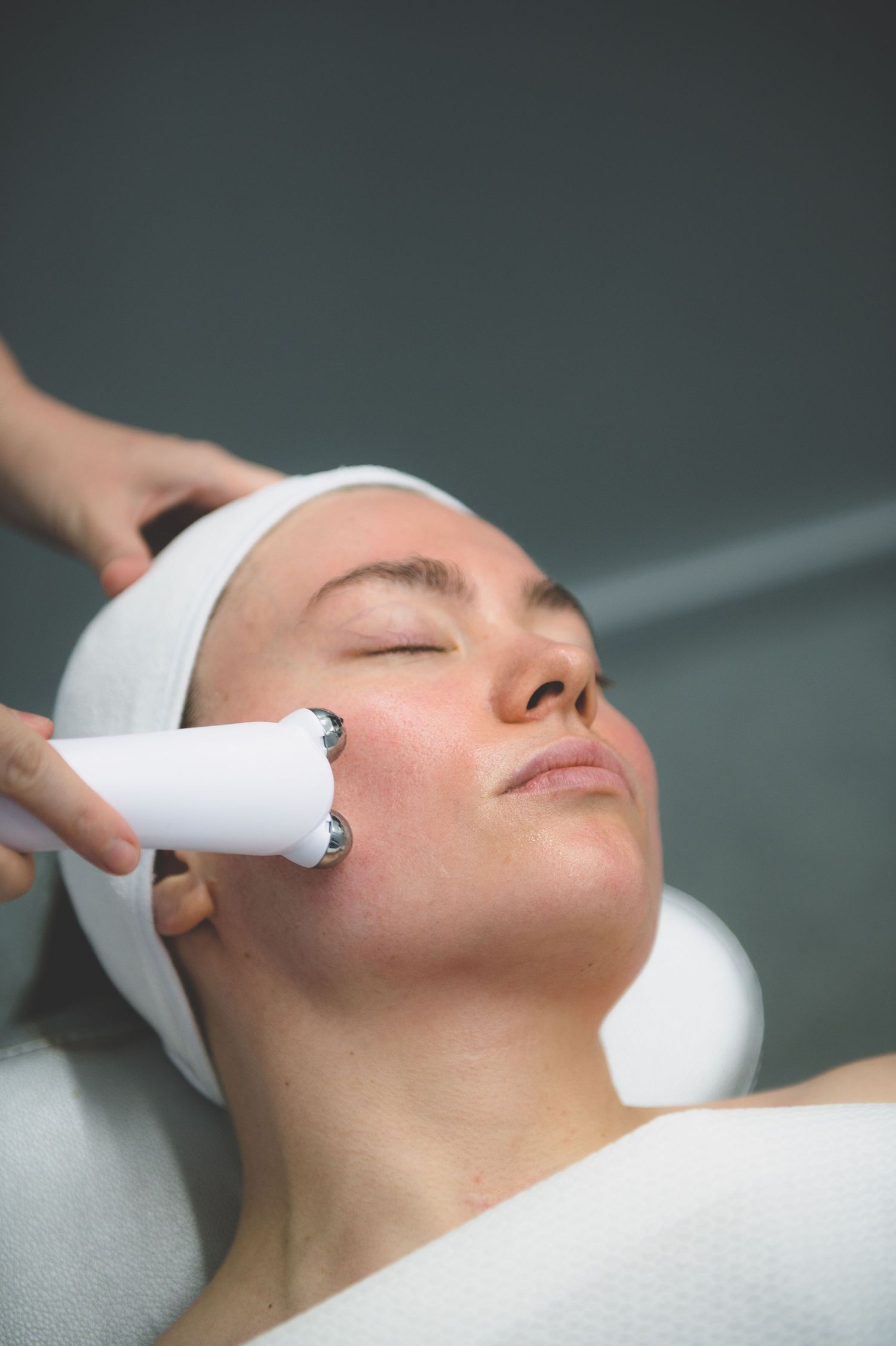Woman receiving a facial treatment with a white device at a beauty spa, wearing a headband with eyes closed.