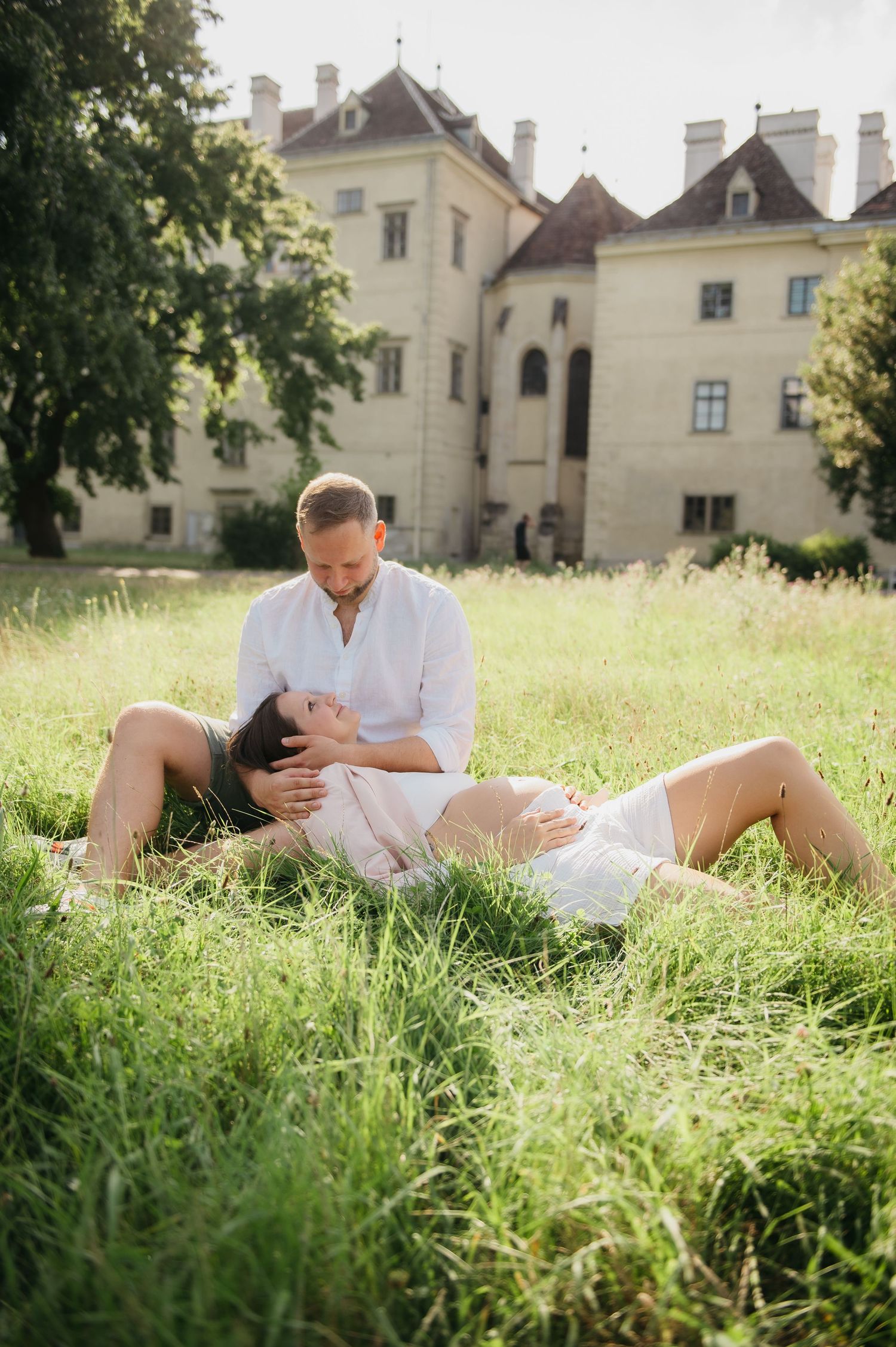 A couple shares a romantic moment in a lush green meadow, with a historic European manor in the background.