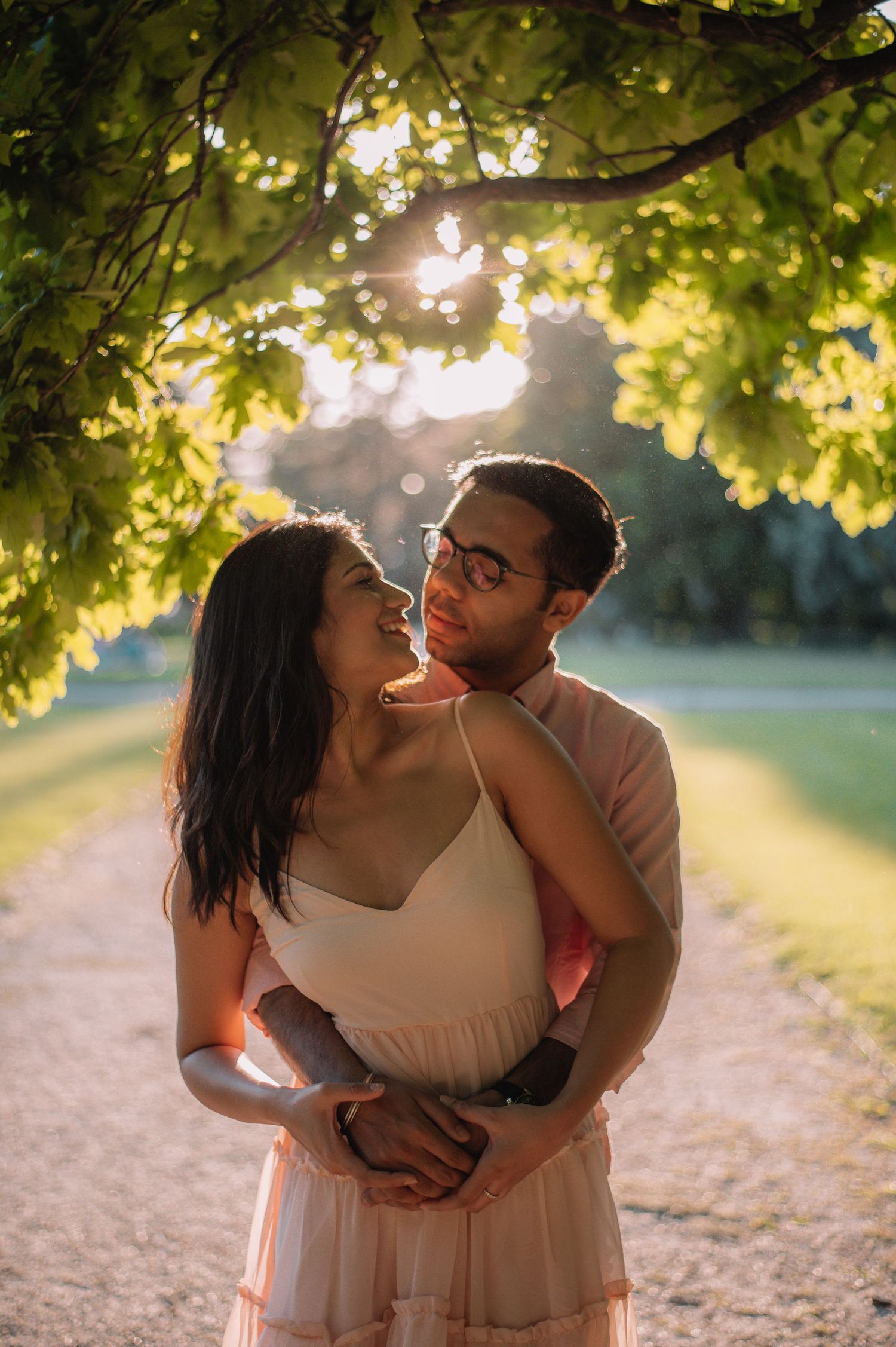 A couple embraces romantically under sunlit green trees on a park path during golden hour sunset.