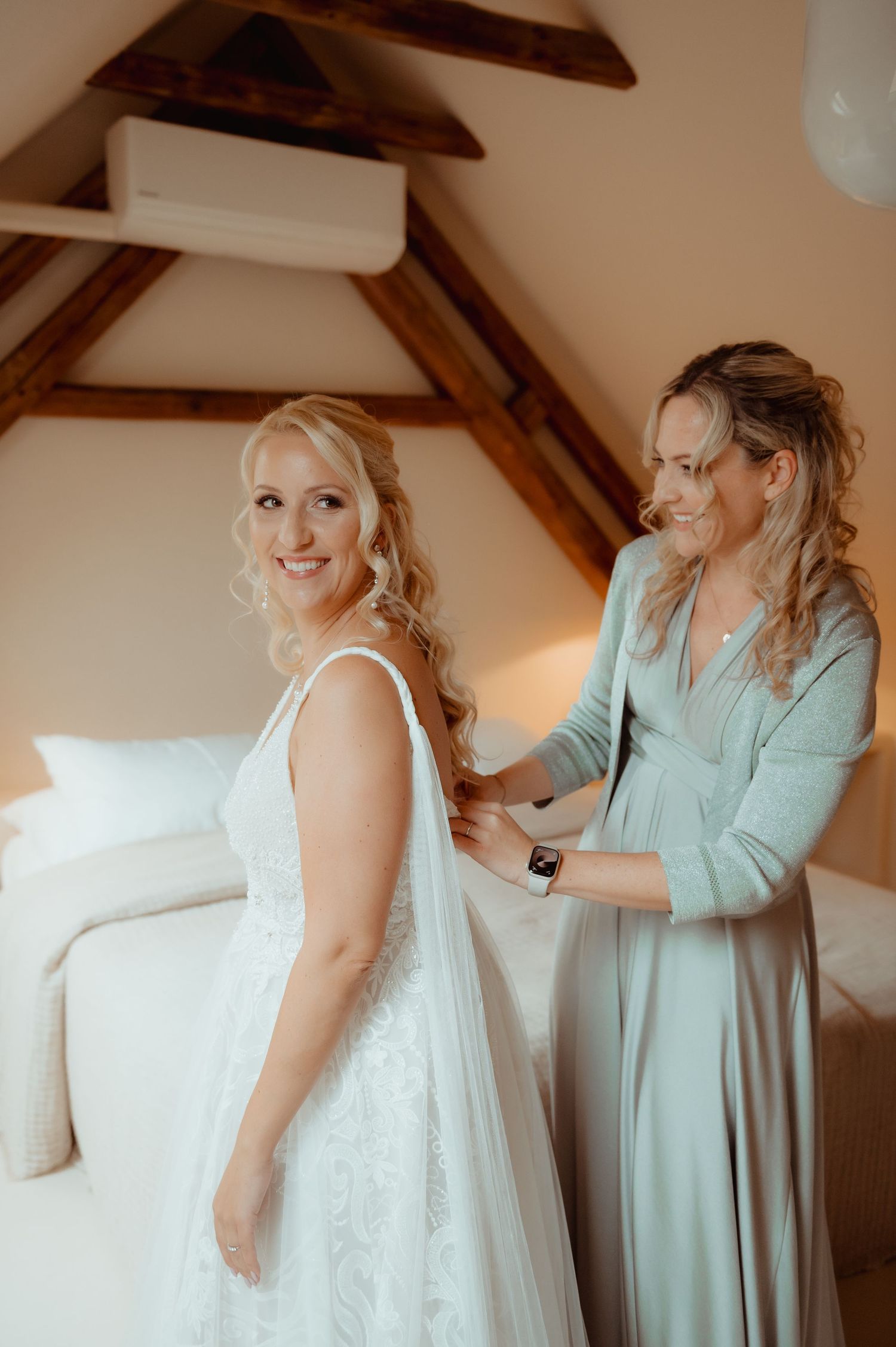 Bride in white lace gown smiles as bridesmaid in sage green dress fastens her veil in a rustic bedroom.