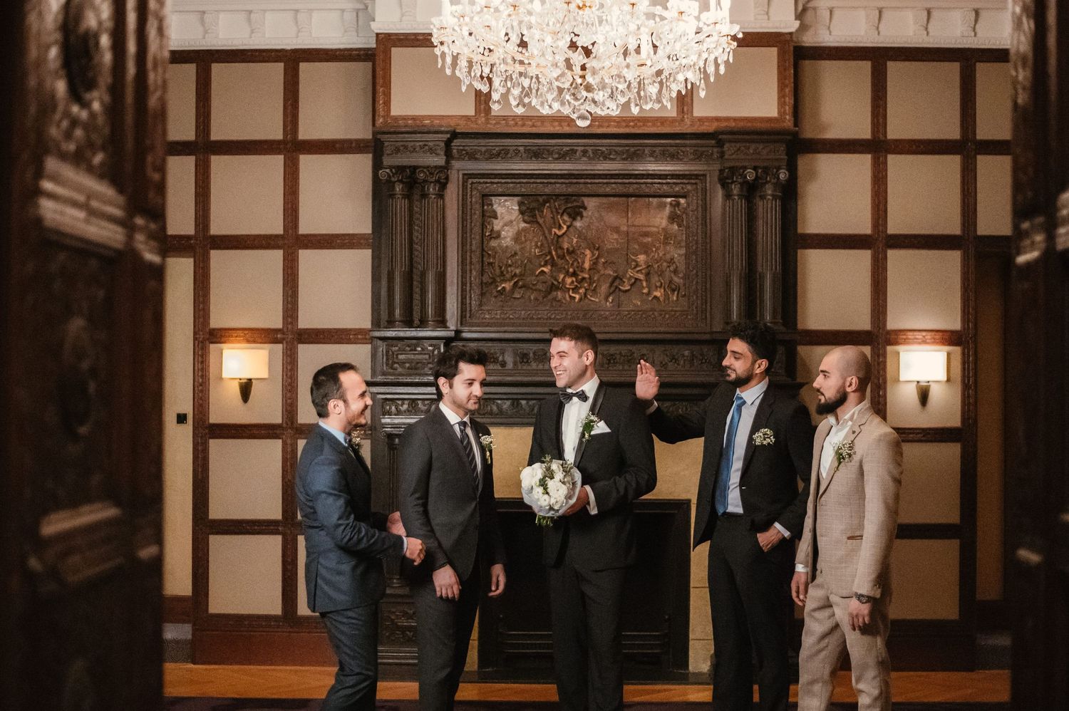 Groom with groomsmen in tuxedos laughing under crystal chandelier in elegant wood-paneled wedding venue.