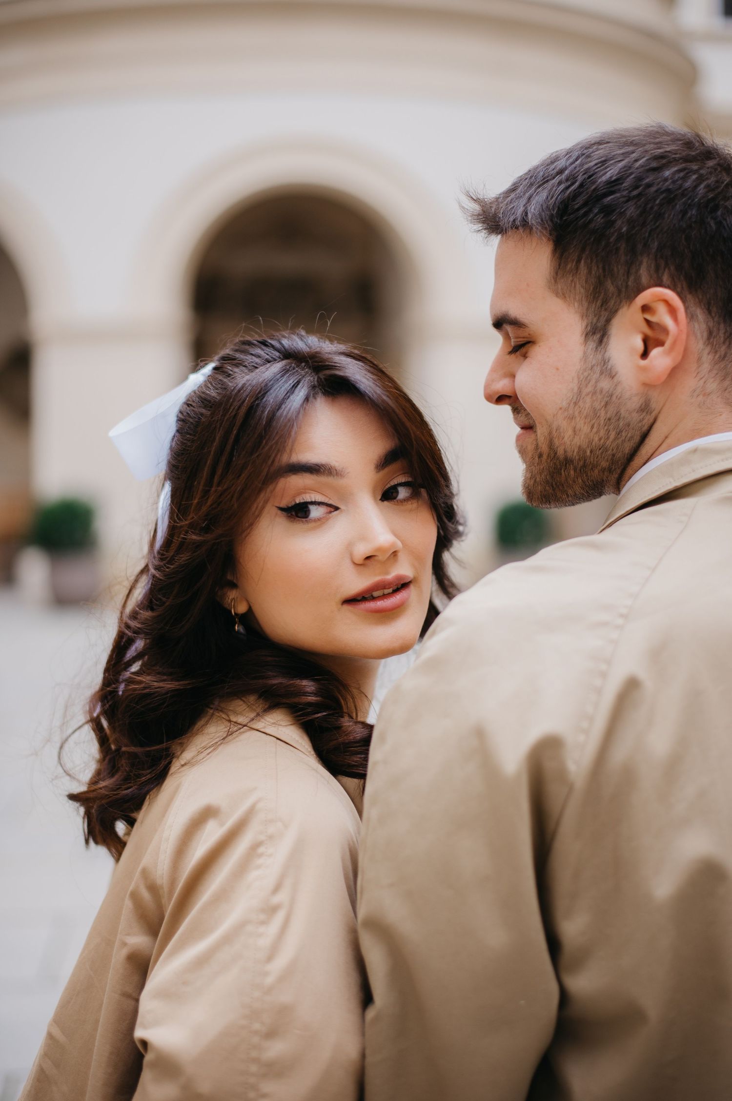 Elegant young woman in beige trench coat gazing softly beside her partner in a classic European courtyard.