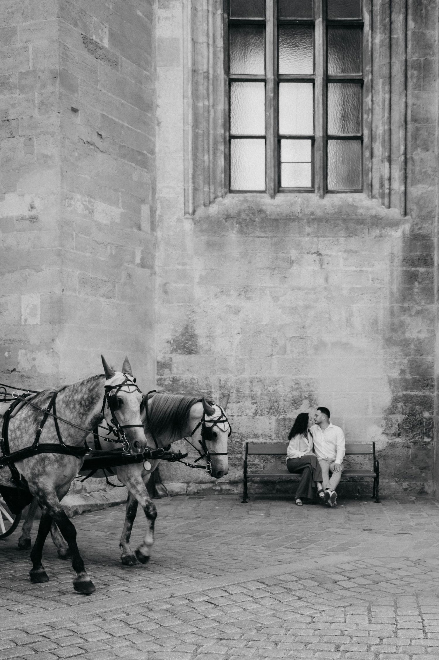 Black and white photo of a romantic couple sitting on a bench beside horse-drawn carriage near historic stone building.