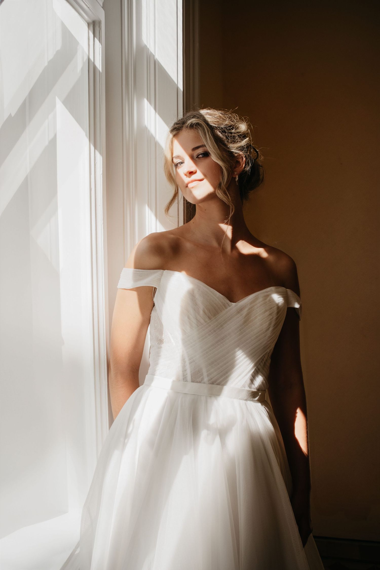 Elegant bride in off-shoulder white ball gown standing by a sunlit window with soft natural light.