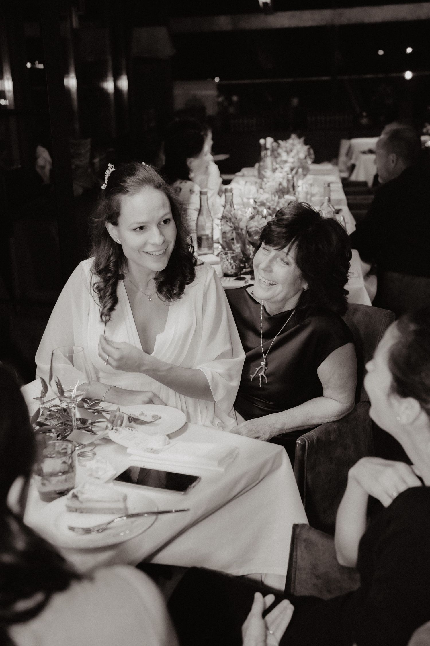 Black and white photo of a smiling bride in white dress chatting with a guest at a wedding reception dinner.