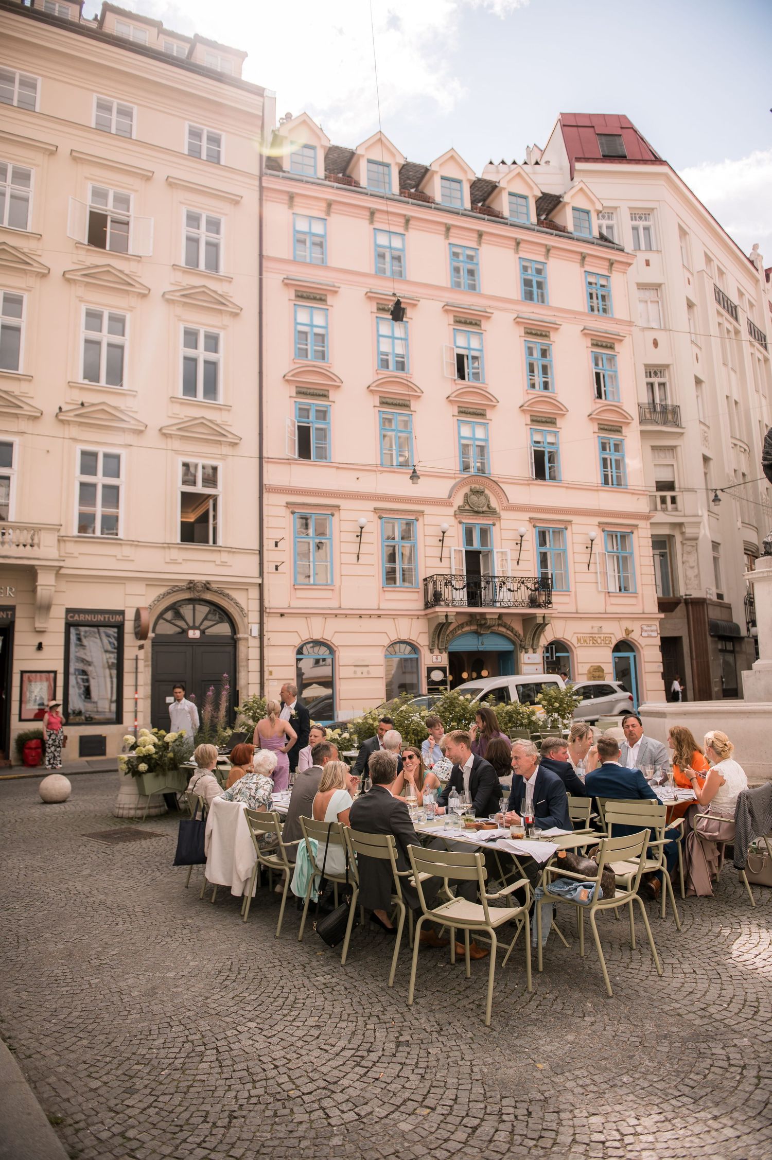 Wedding guests dining at long outdoor table in cobblestone square before grand European building facade.