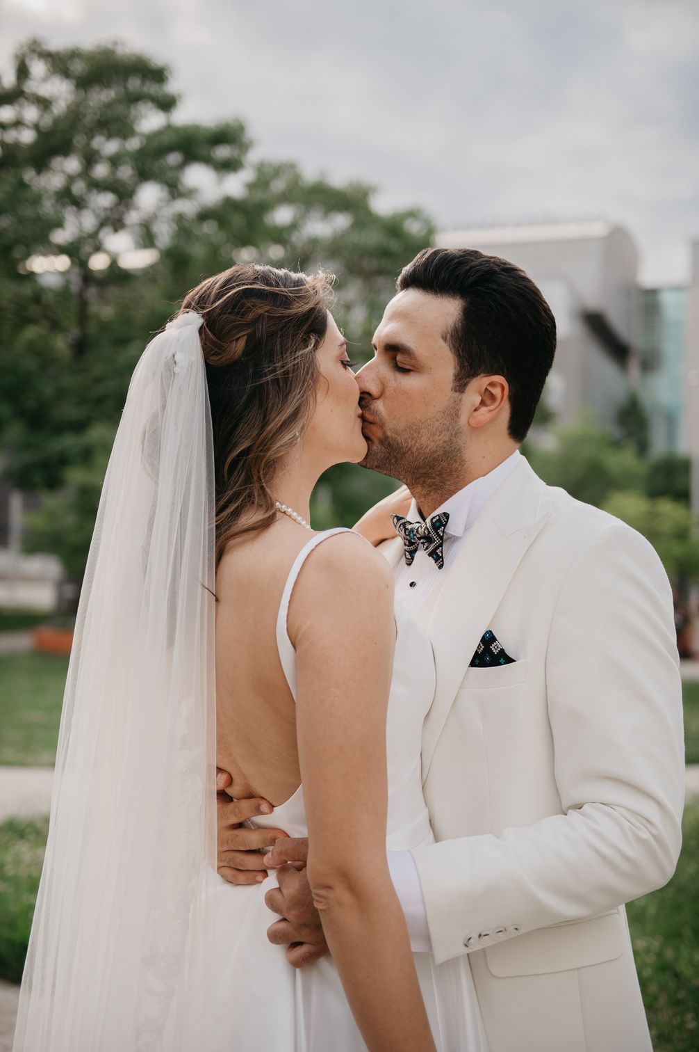 Bride and groom sharing a tender kiss outdoors, groom in white suit with floral bow tie, bride in veil.