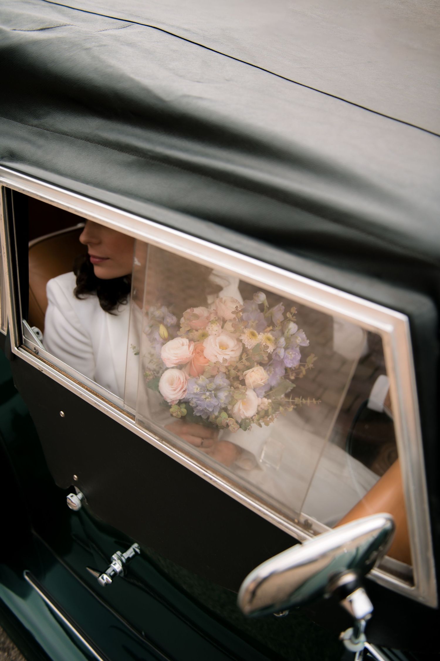 Bride in white dress holding bouquet of pink and white flowers looks out vintage car window.
