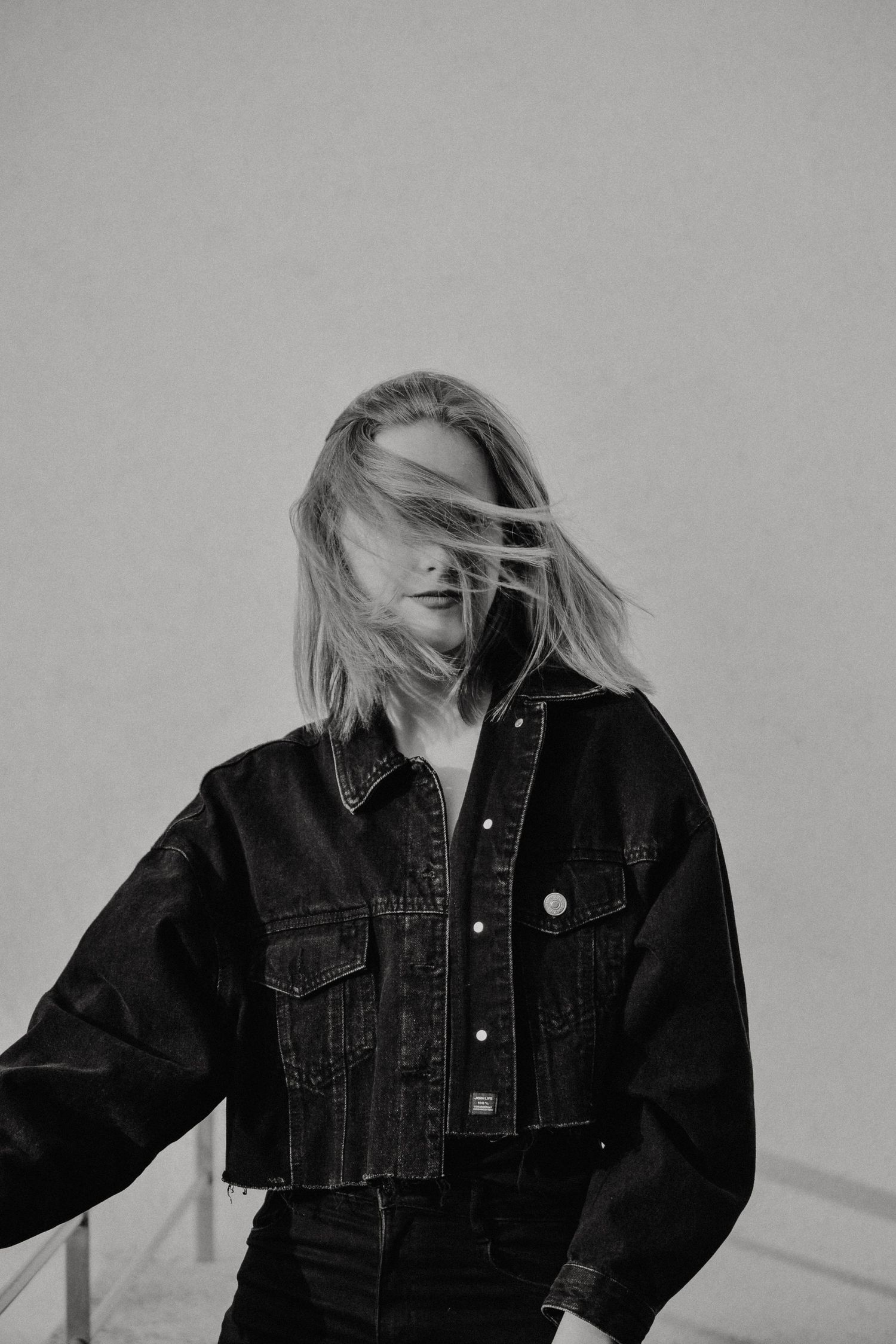 Black and white photo of a blonde woman in a denim jacket with hair blowing across her face.