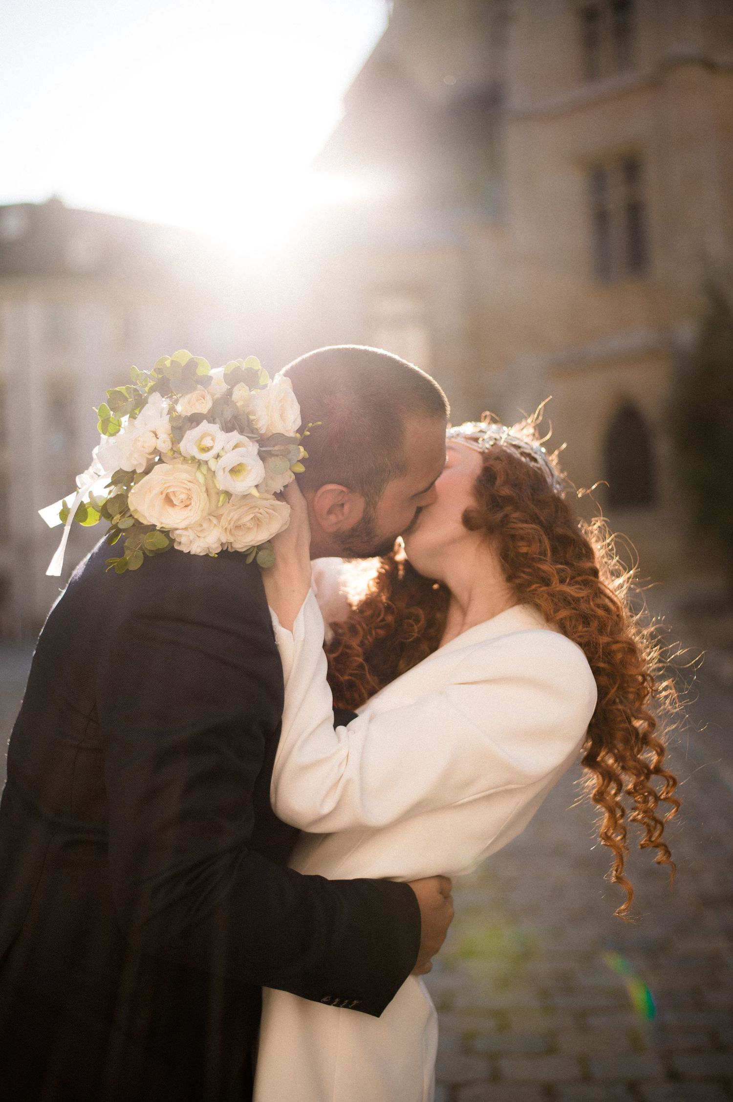 Bride and groom share a passionate kiss backlit by golden sunlight on a cobblestone street.