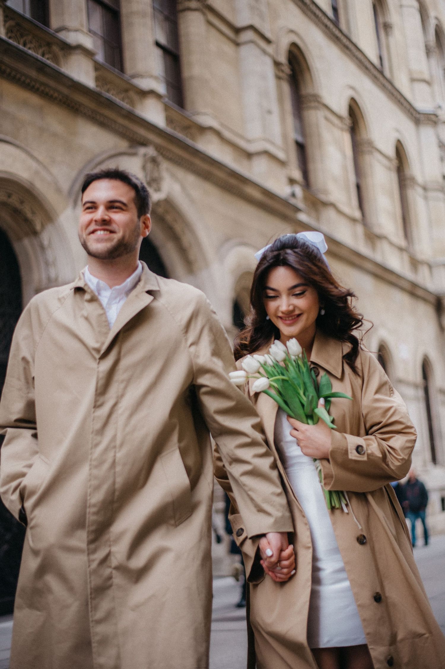 Happy couple in matching beige trench coats walking hand in hand while woman holds white tulips near historic building.