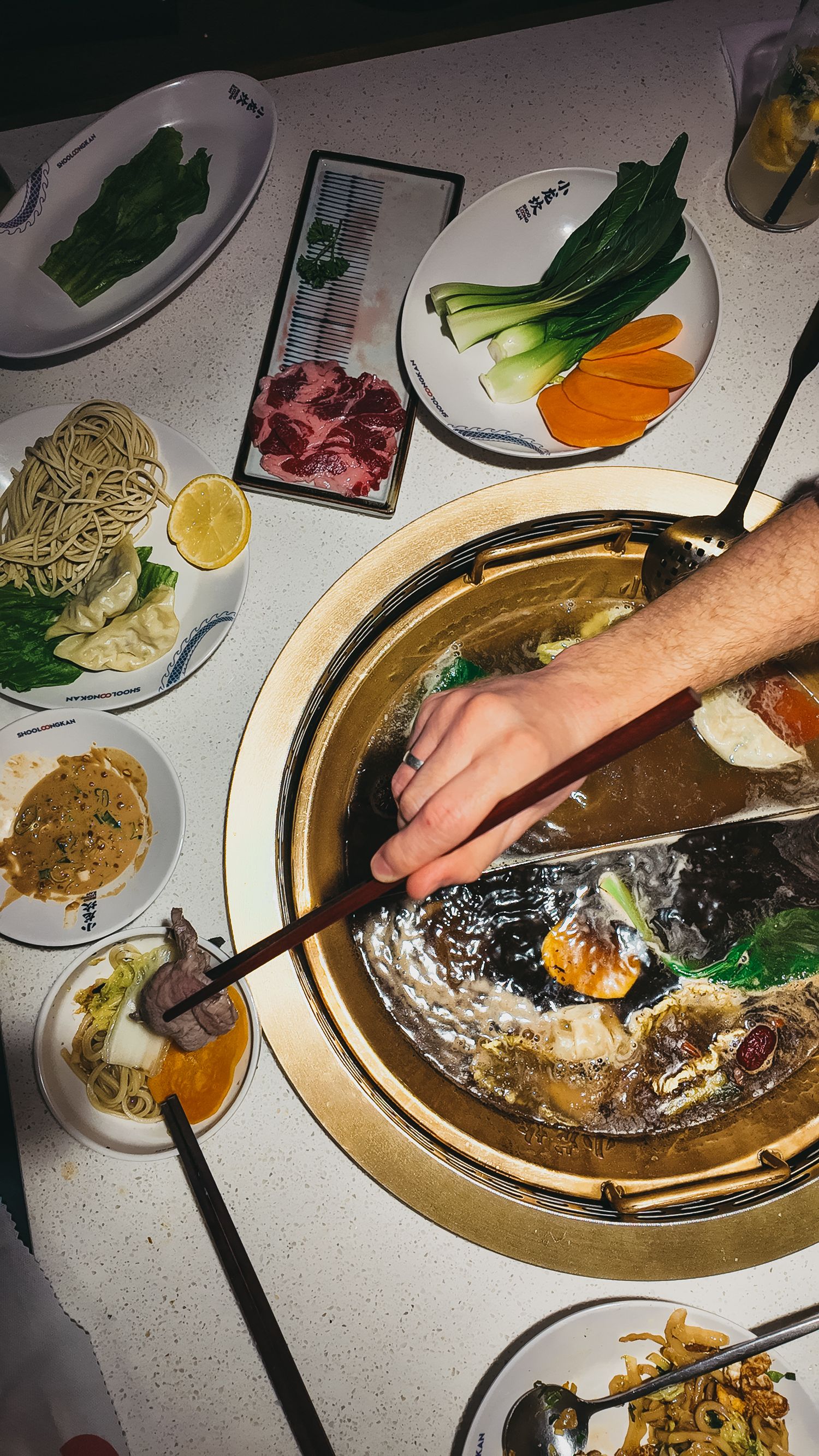 A hand using chopsticks to cook ingredients in a hot pot surrounded by raw meats, noodles, and vegetables.