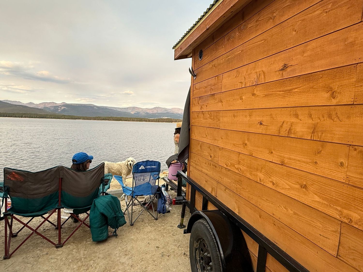 Wooden cabin structures along a lakeside with mountain views.