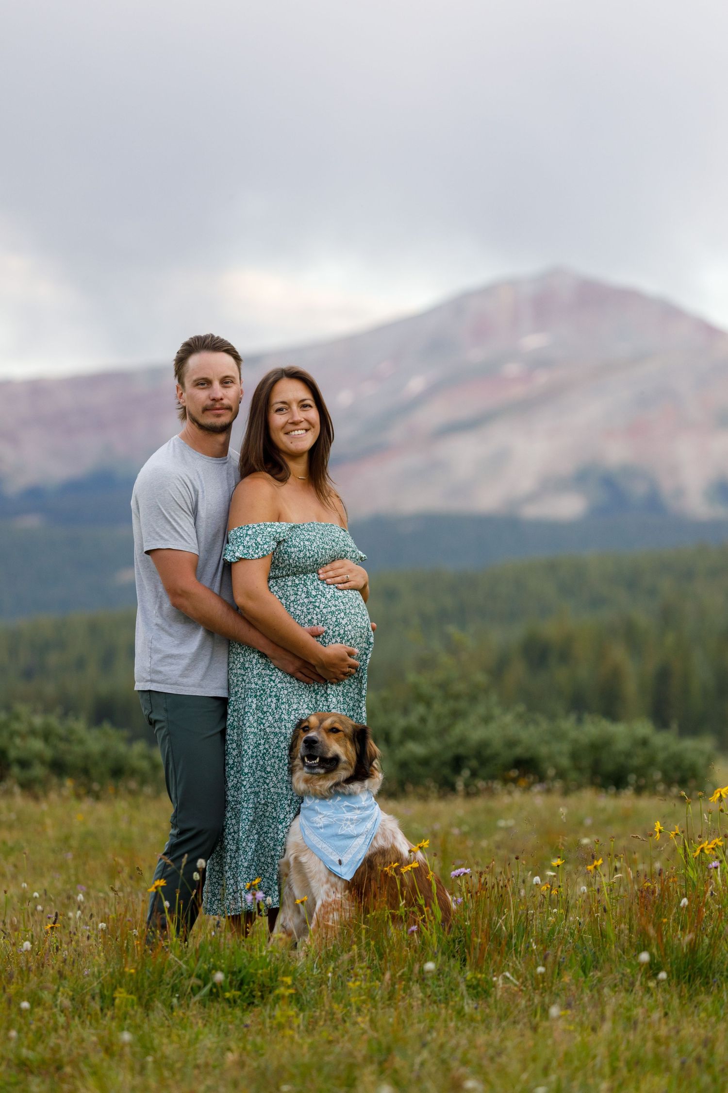Family with dog posing in mountain meadow with peaks in background during summer.