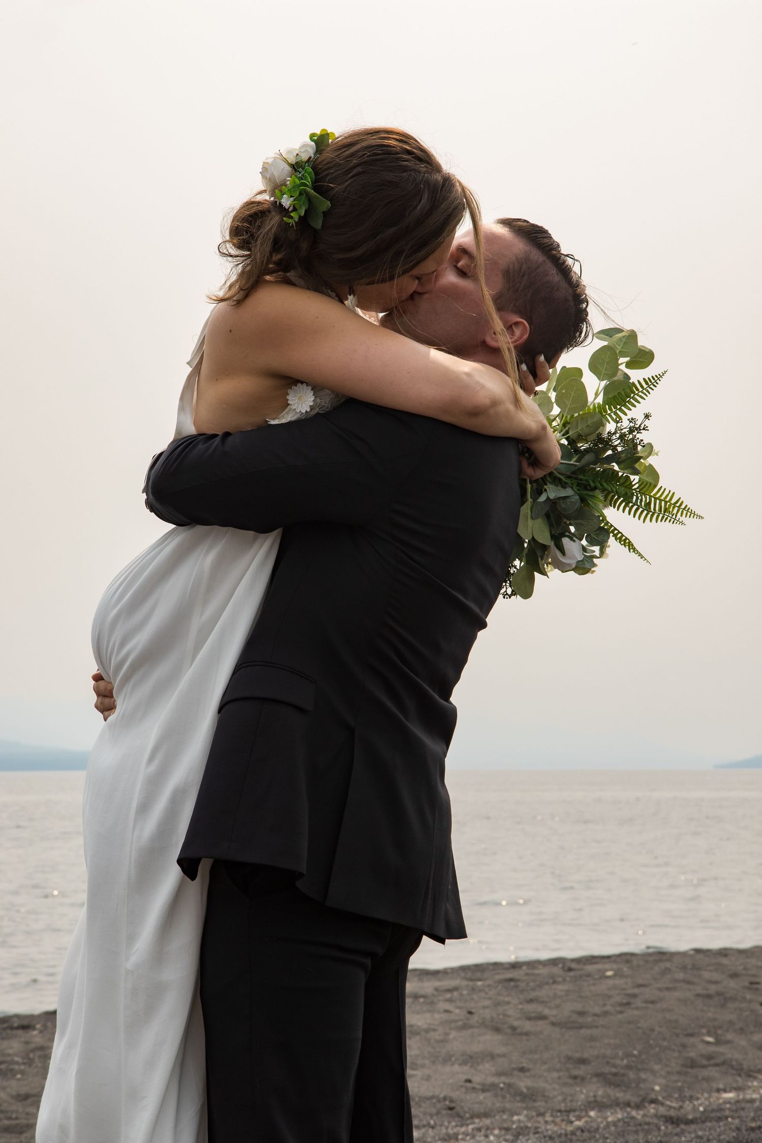 Couple embracing on beach with greenery bouquet against ocean backdrop.