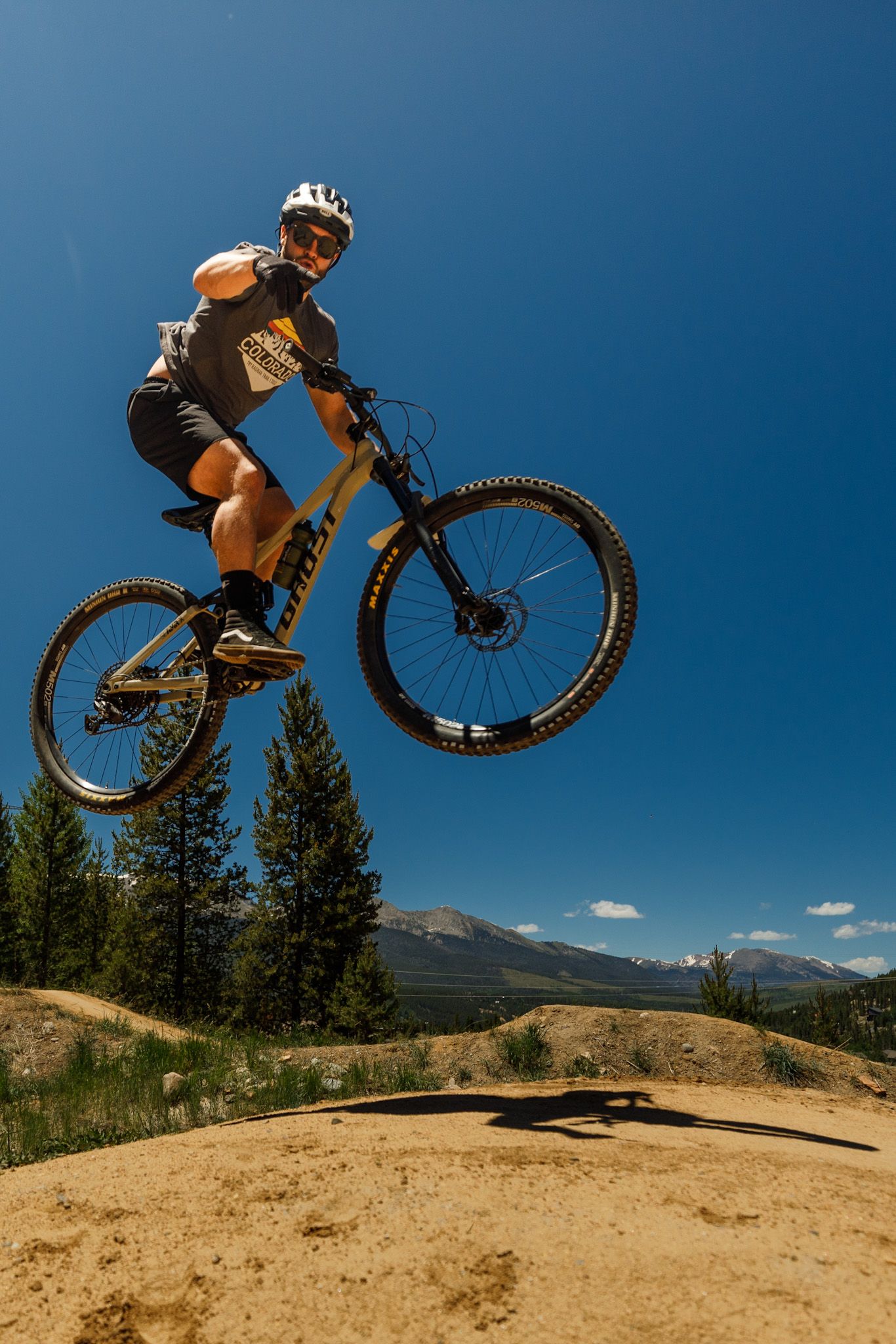 Mountain biker performing aerial jumps on dirt trails against blue sky and mountain backdrop.