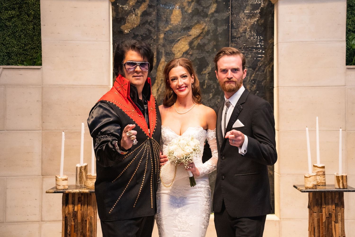 A wedding party poses together in formal attire against a stone wall backdrop with candlelit sconces.