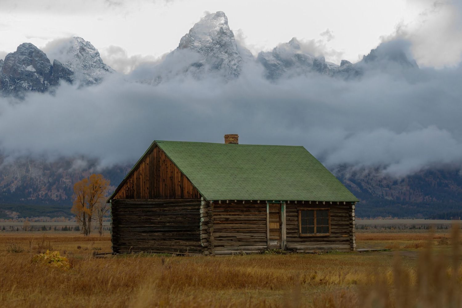 A rustic log cabin with green roof sits beneath misty Grand Teton mountains in autumn.
