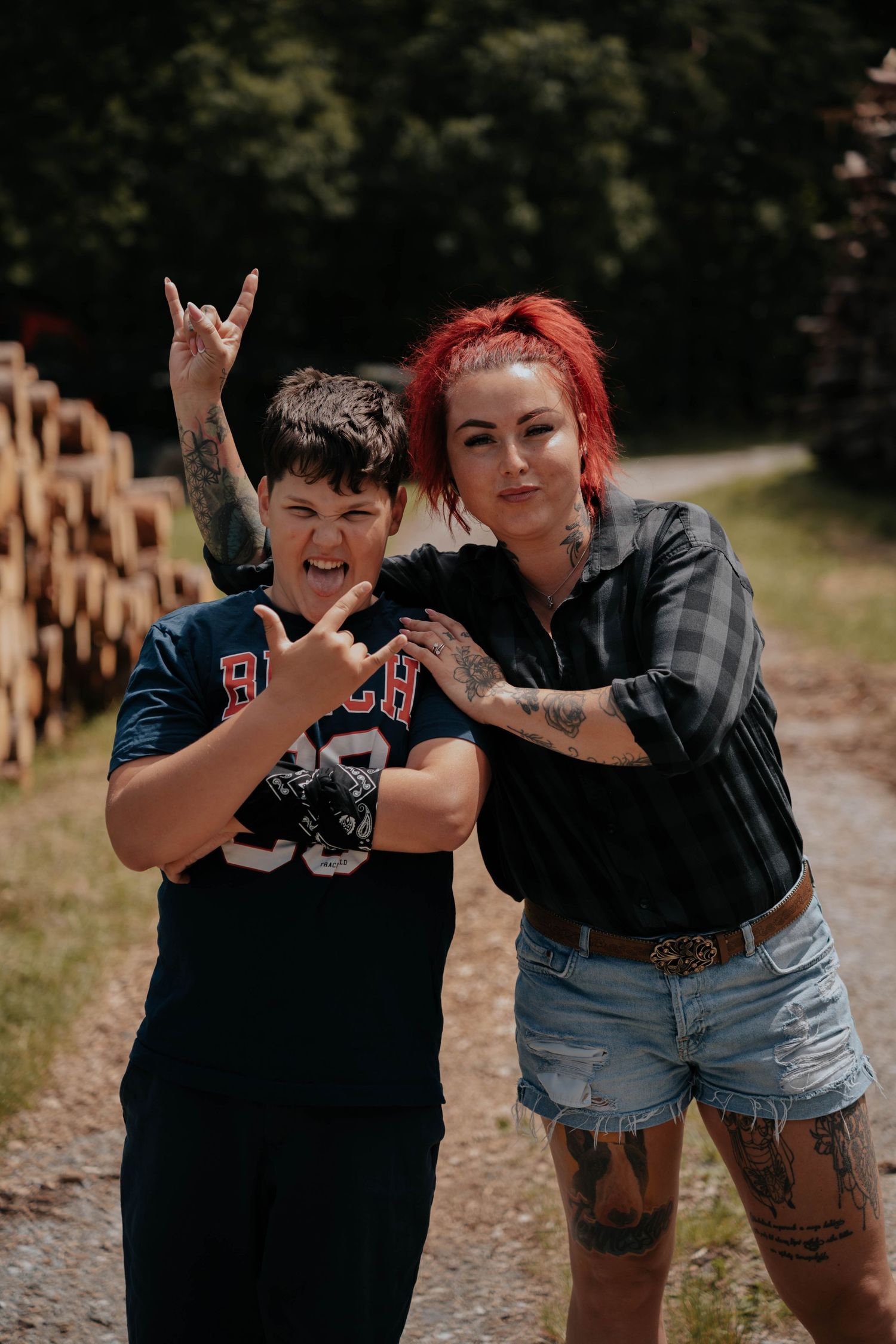 Two people posing outdoors making rock on hand gestures, one with red hair and tattoos in plaid shirt and denim shorts.