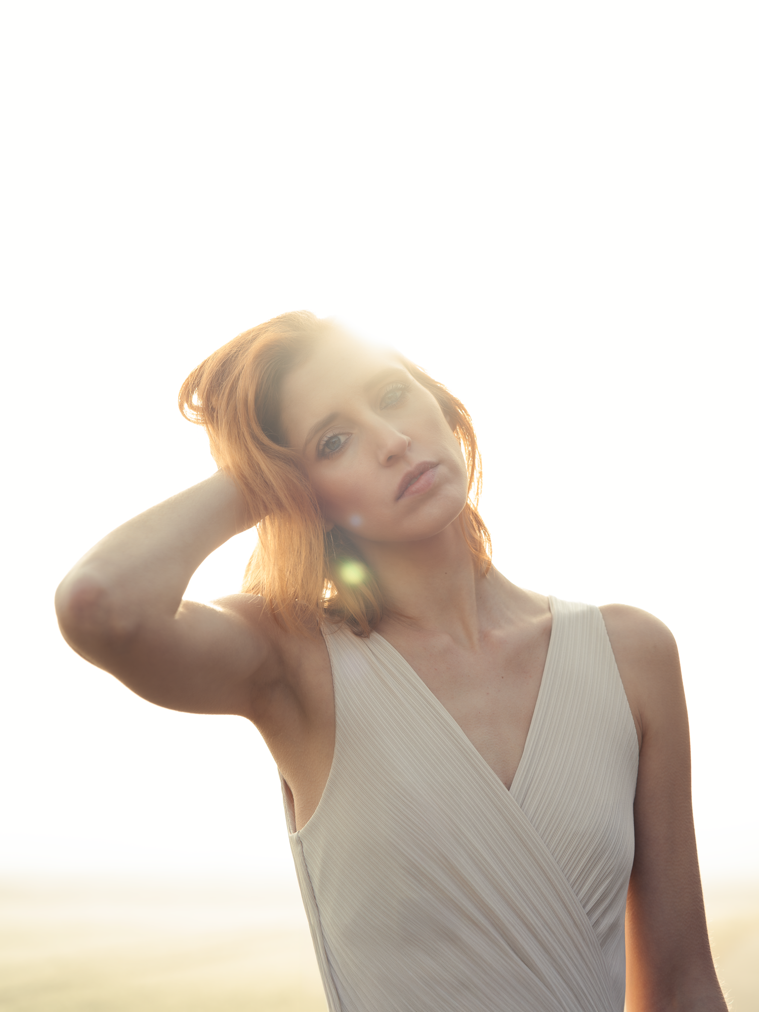 A young woman in a white wrap dress poses with hand in hair, bathed in golden backlit sunlight.