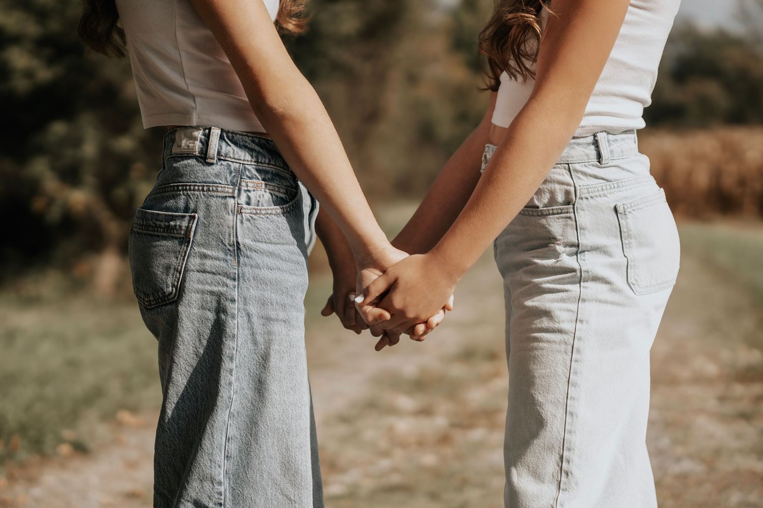Two women holding hands outdoors in jeans, symbolizing friendship or love in a natural setting.