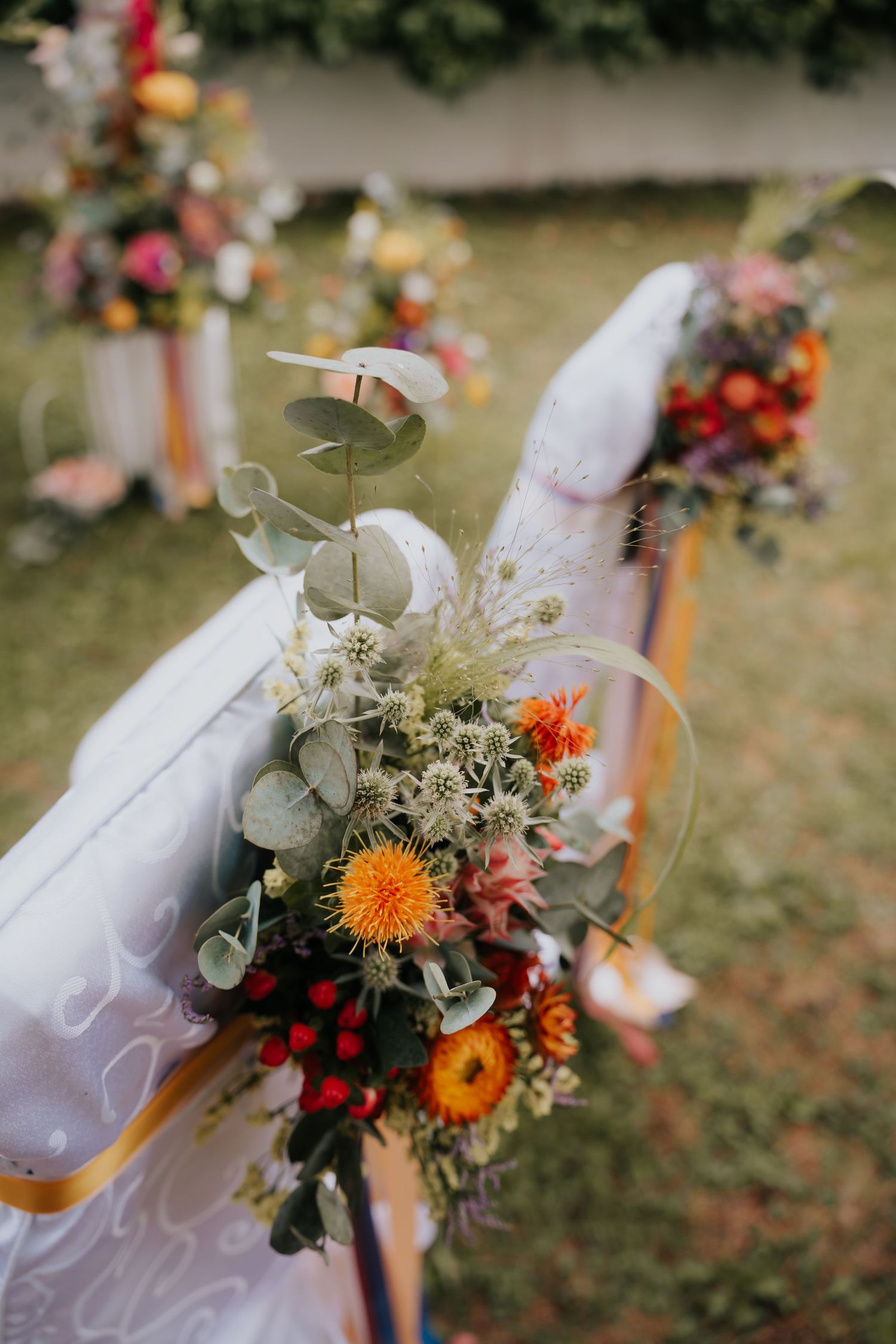 Colorful floral wedding aisle decorations with orange, red, and white blooms on white chairs outdoors.