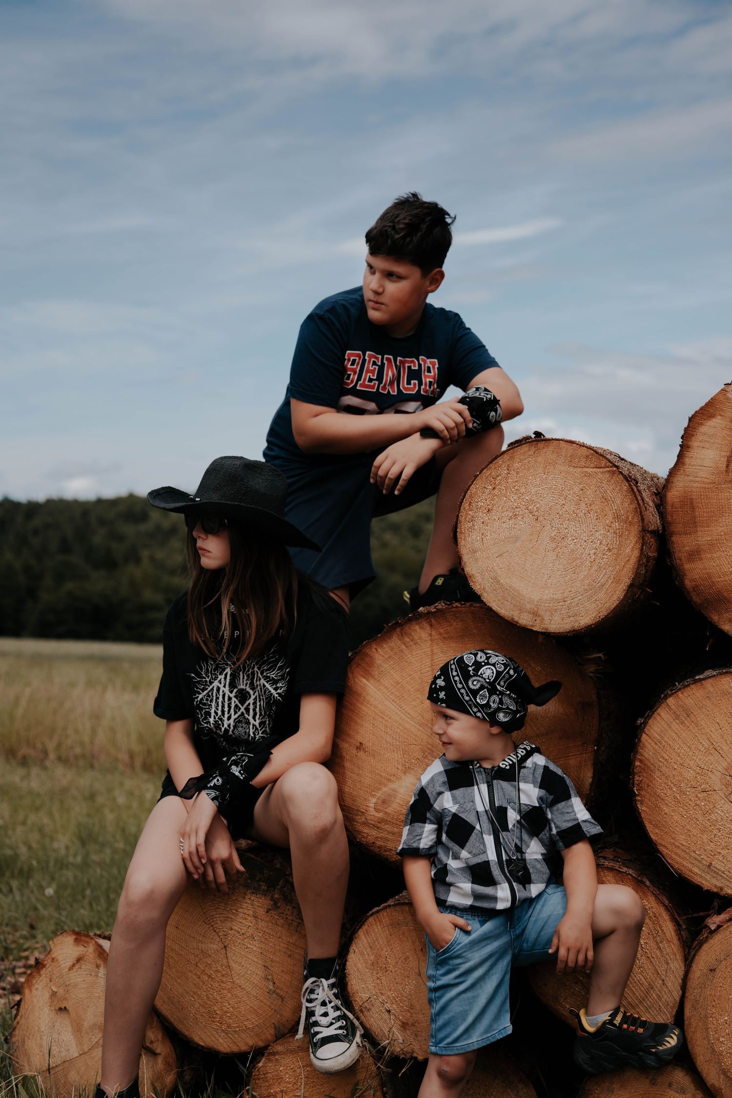 Three kids posing on a stack of logs in a field, wearing casual outfits including a cowboy hat and bandana.