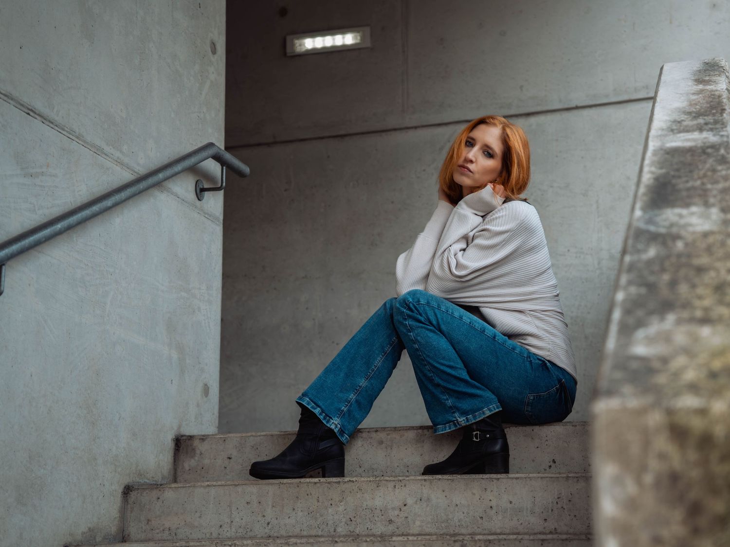 A red-haired woman in jeans and white sweater sits thoughtfully on concrete stairs wearing black ankle boots.