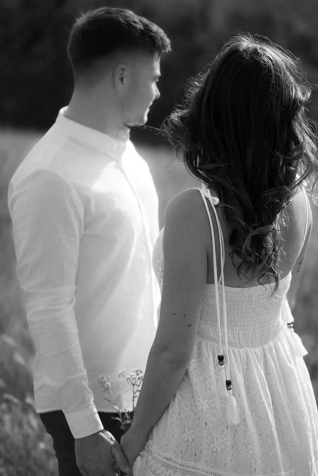 Black and white photo of a couple holding hands, woman in white dress with long curly hair, man in white shirt.