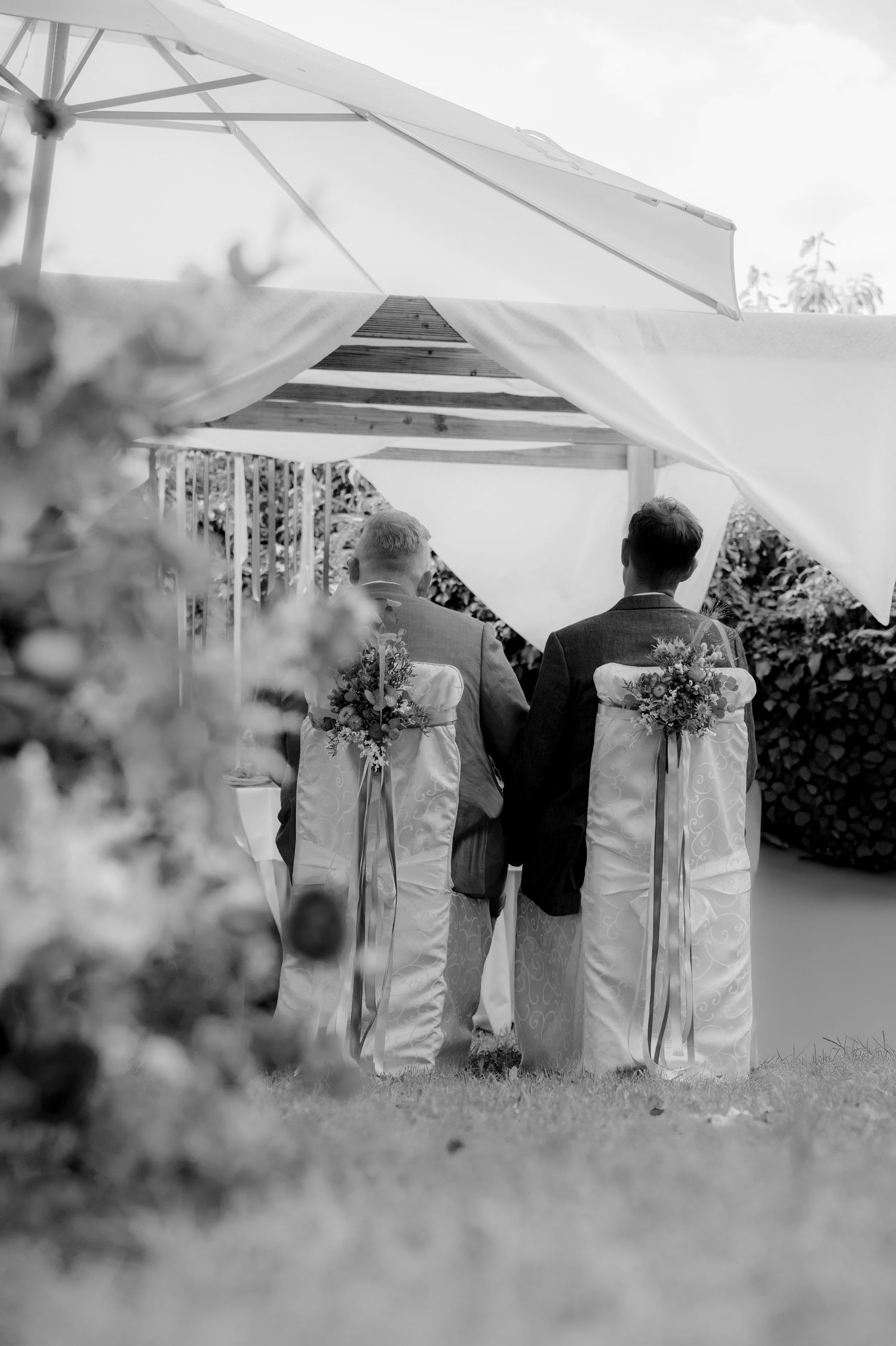 Black and white photo of two grooms standing at outdoor wedding altar with floral bouquets and white chair sashes.