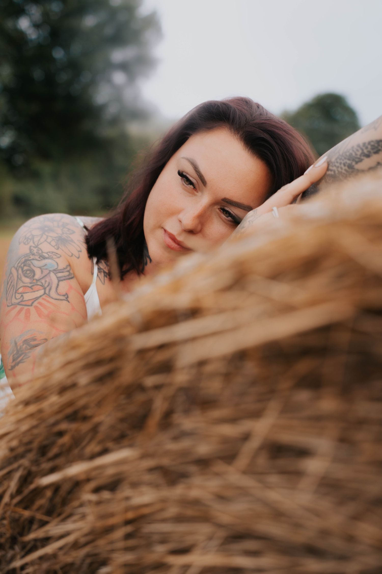Close-up of a tattooed brunette woman leaning against a rustic hay bale in a serene outdoor setting.
