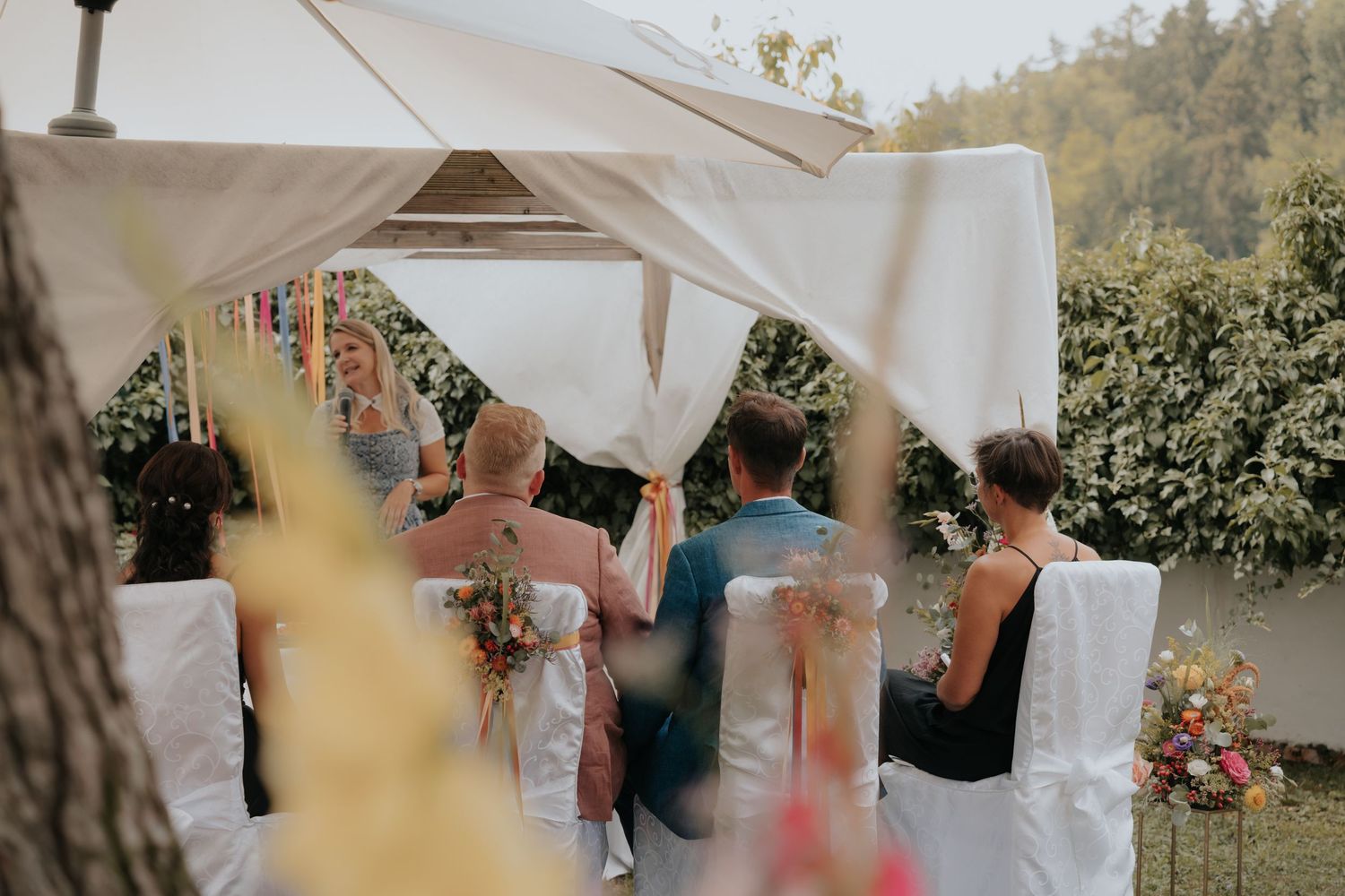 Outdoor wedding ceremony under a white canopy with guests seated in white chairs surrounded by lush greenery.