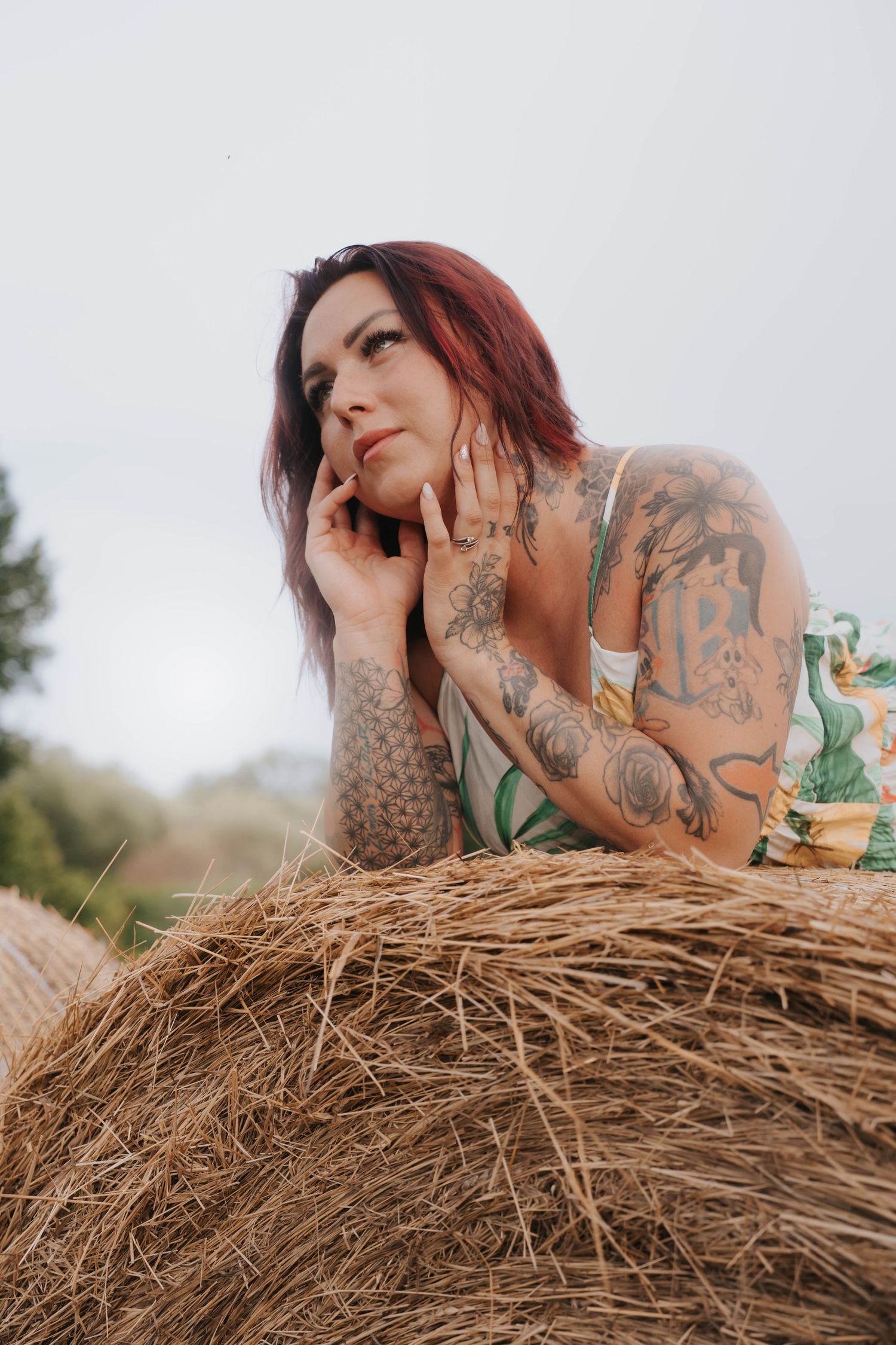 Tattooed woman with red hair resting on a hay bale outdoors, gazing upward in a thoughtful pose.