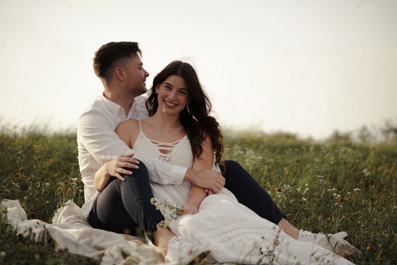 Couple sitting together in a sunlit field, woman in white dress smiling warmly, man embracing her from behind.