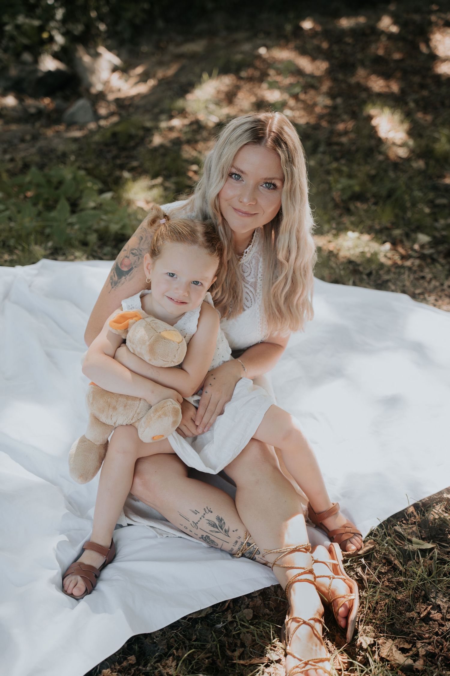Blonde mother with tattoos sits on white blanket outdoors, hugging young red-haired girl holding a stuffed animal toy.