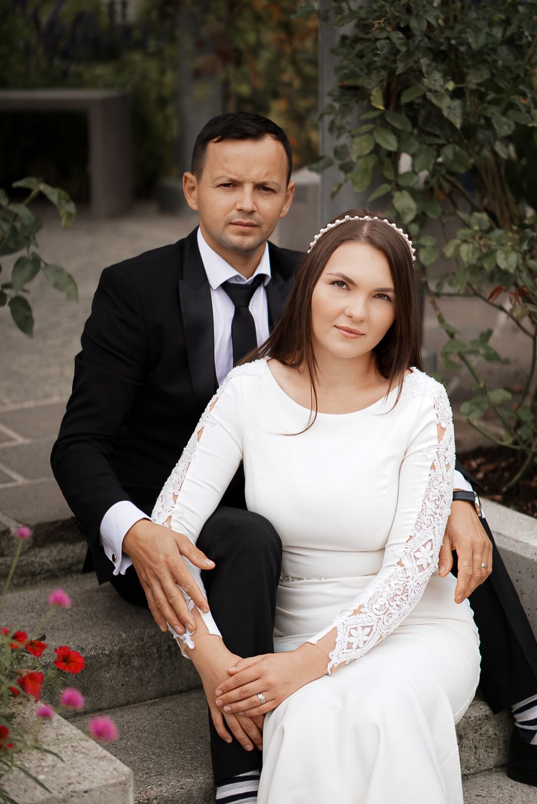 A couple in wedding attire sits on stone steps surrounded by greenery and red flowers, the bride in white lace and groom in black suit.