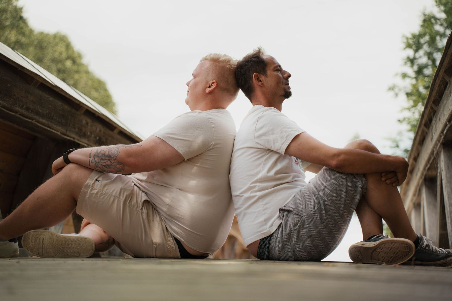 Two men in white t-shirts and shorts sit back-to-back on a wooden deck outdoors.