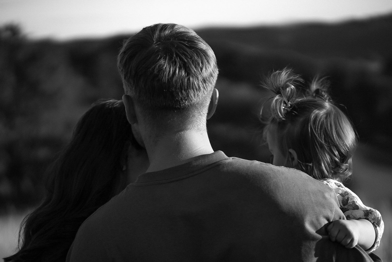 Black and white photo of a couple embracing from behind, gazing at a scenic outdoor landscape together.