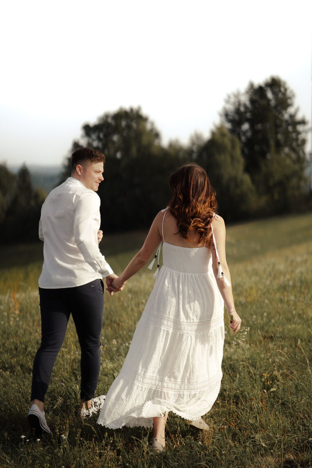 Couple holding hands walking through green meadow, woman in white dress, man in white shirt and dark pants, trees in background.
