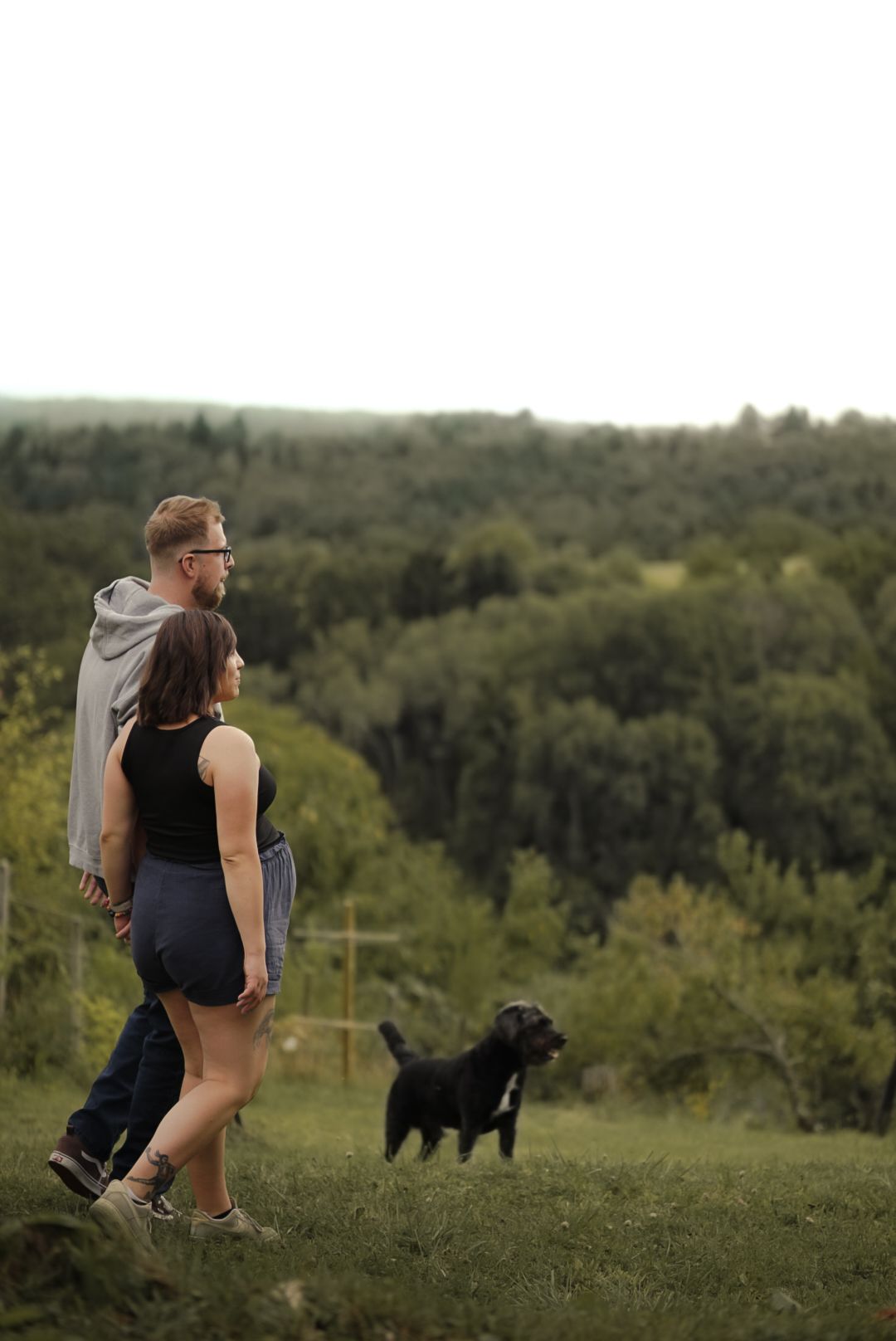Couple with black dog enjoying scenic green forest view from hilltop on cloudy day.