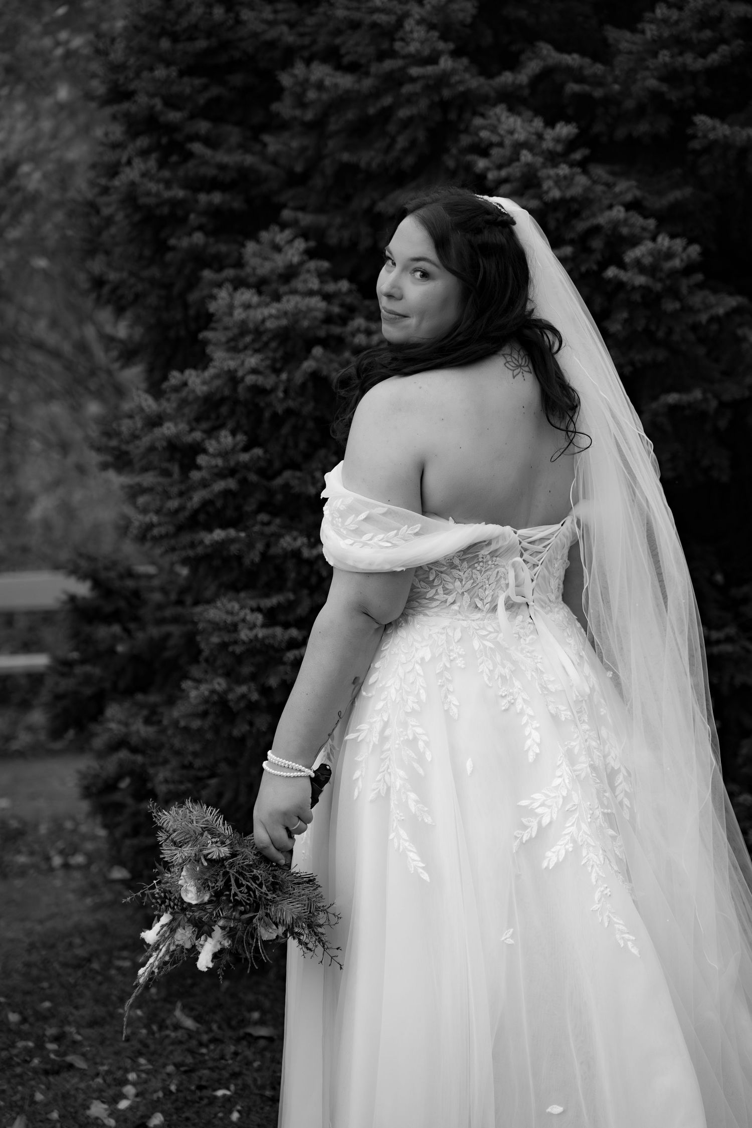 Black and white photo of a bride in an off-shoulder lace gown and veil, holding a bouquet, looking back over her shoulder.