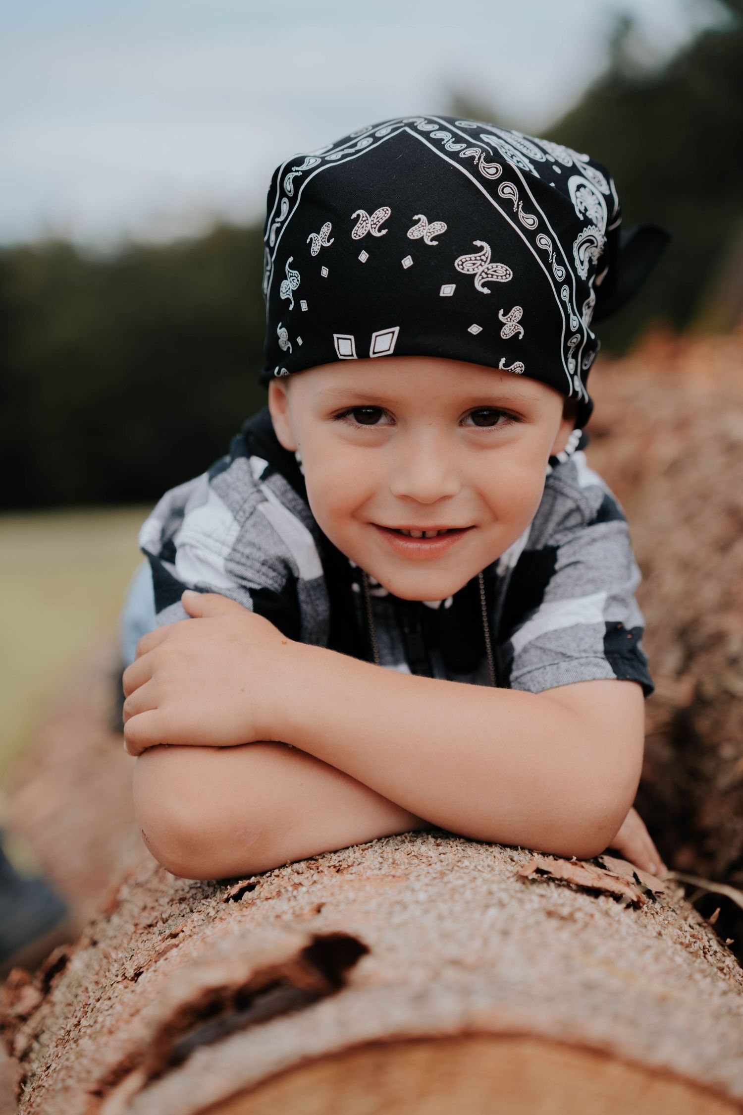 Young boy wearing a black bandana and plaid shirt, smiling while leaning on a log outdoors.