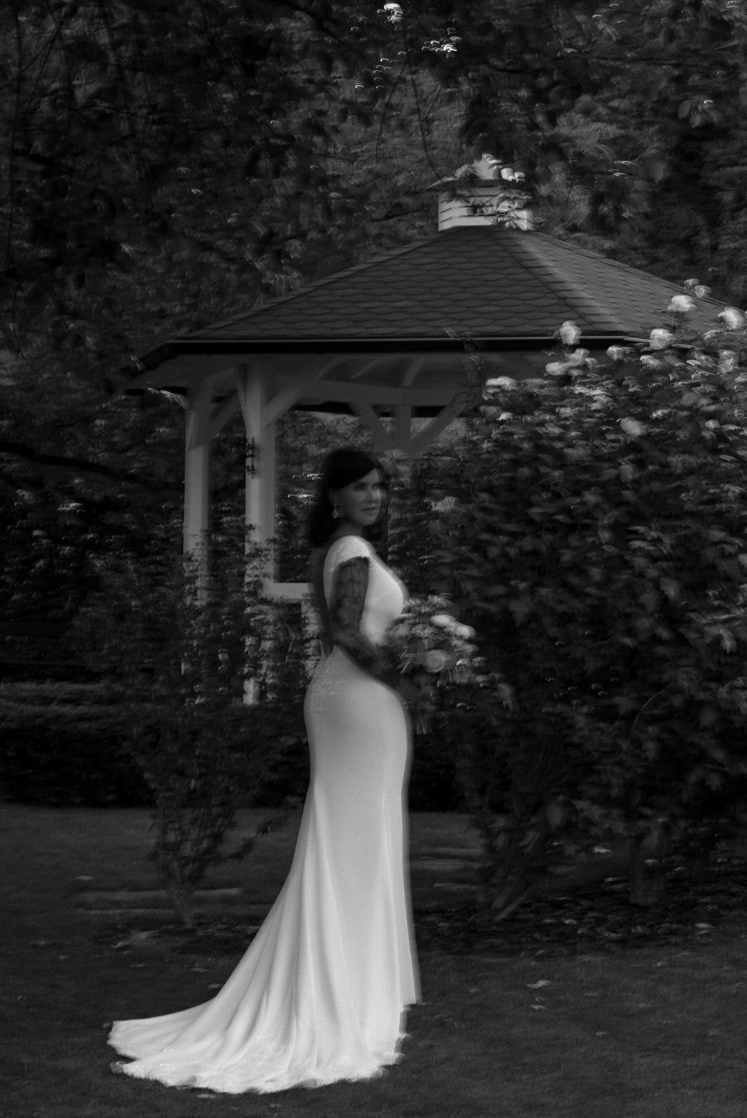 Bride in elegant white gown poses in garden with gazebo and roses in black and white wedding portrait.