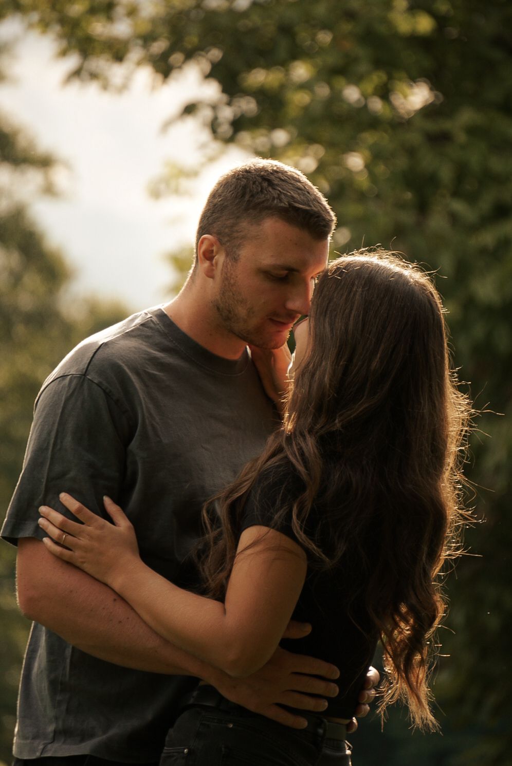 A couple shares a romantic moment, embracing and almost kissing outdoors with soft golden light and lush green trees behind them.